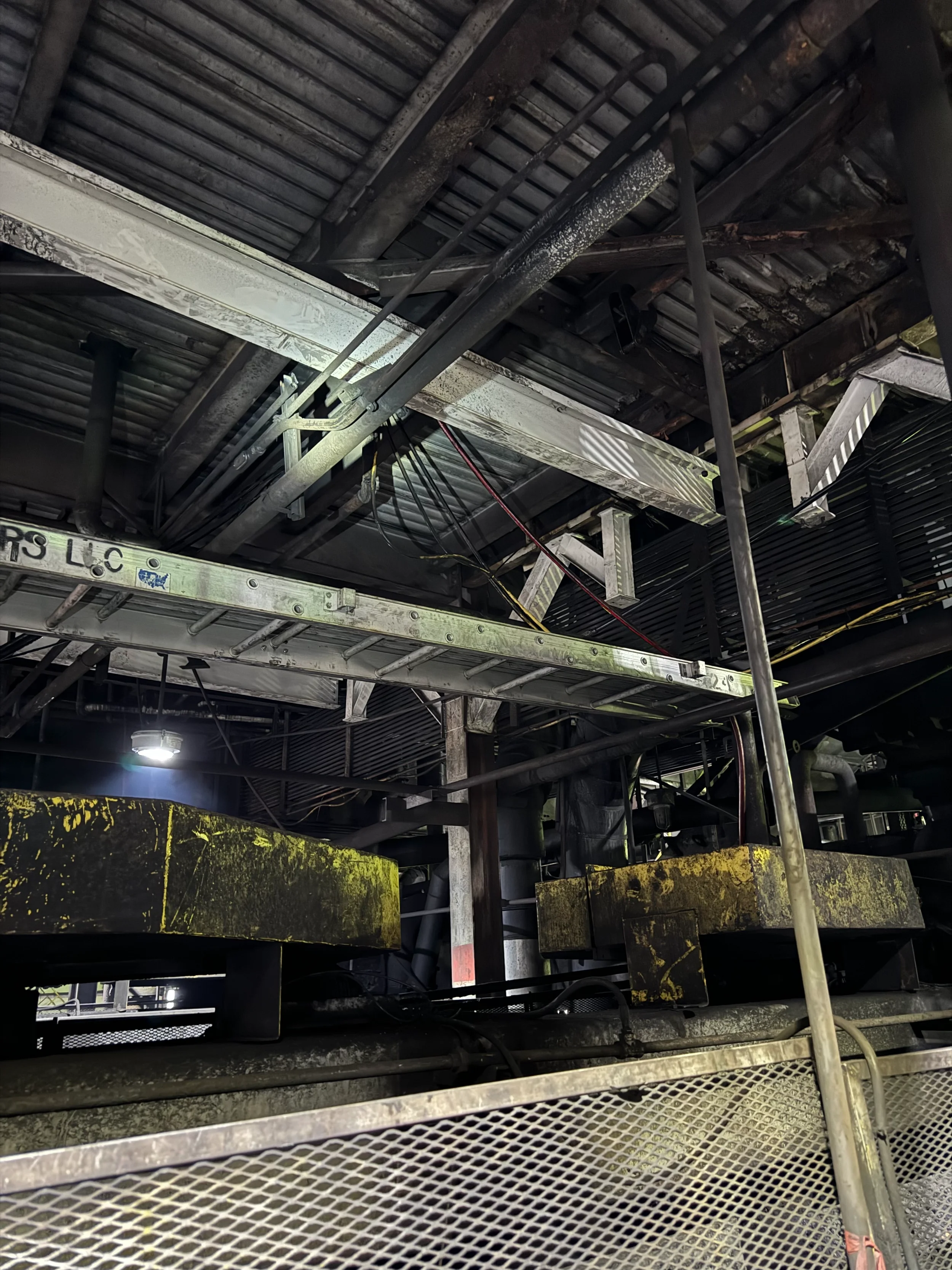 A view of industrial machinery and structural elements in a factory or warehouse setting, including metal pipes, a ladder, and various metal surfaces with signs of corrosion and wear.