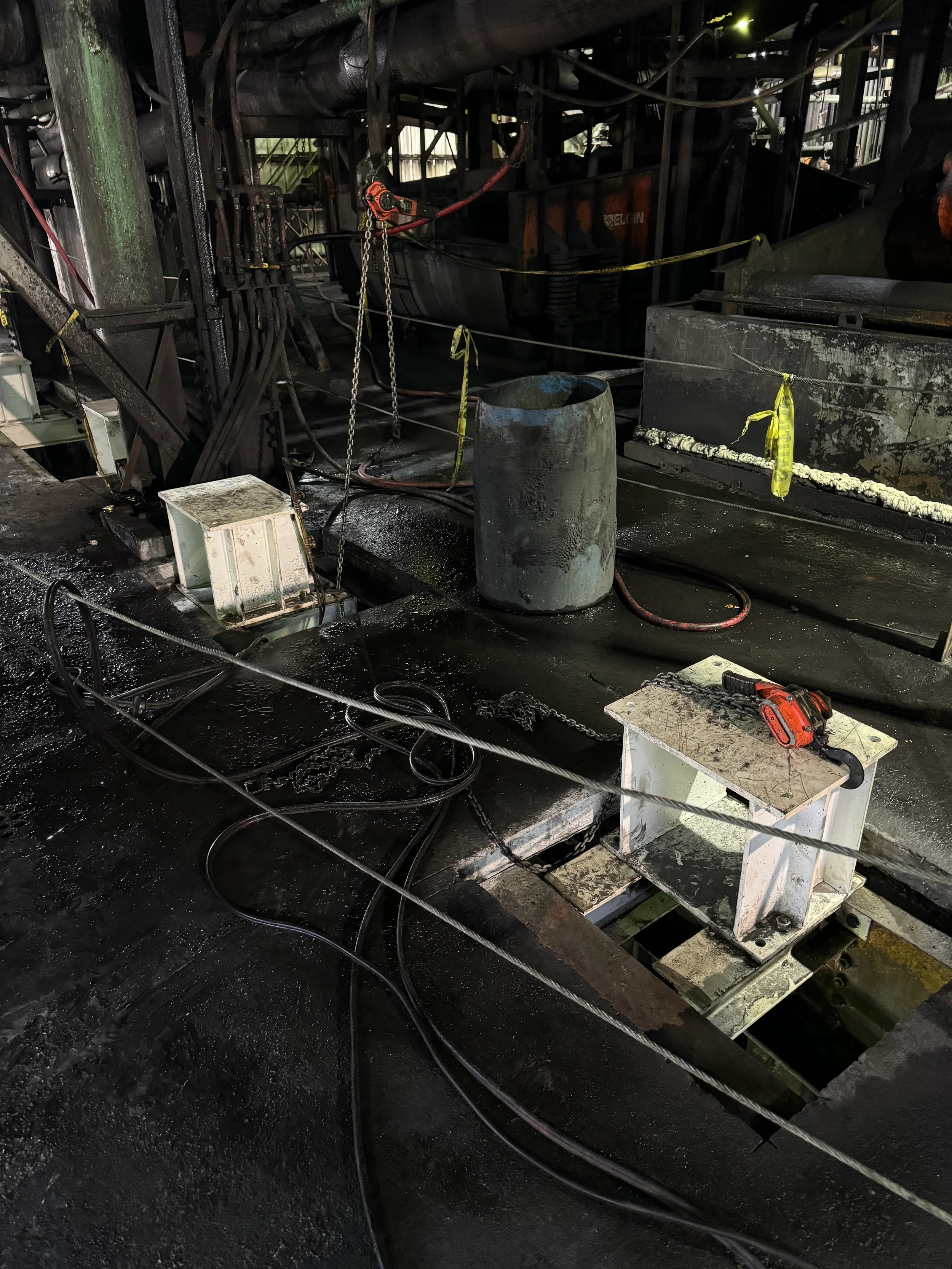 Industrial scene with machinery, cables, chains, and a black, oily floor. There is a cylindrical metal object, a white table, and yellow caution tape.