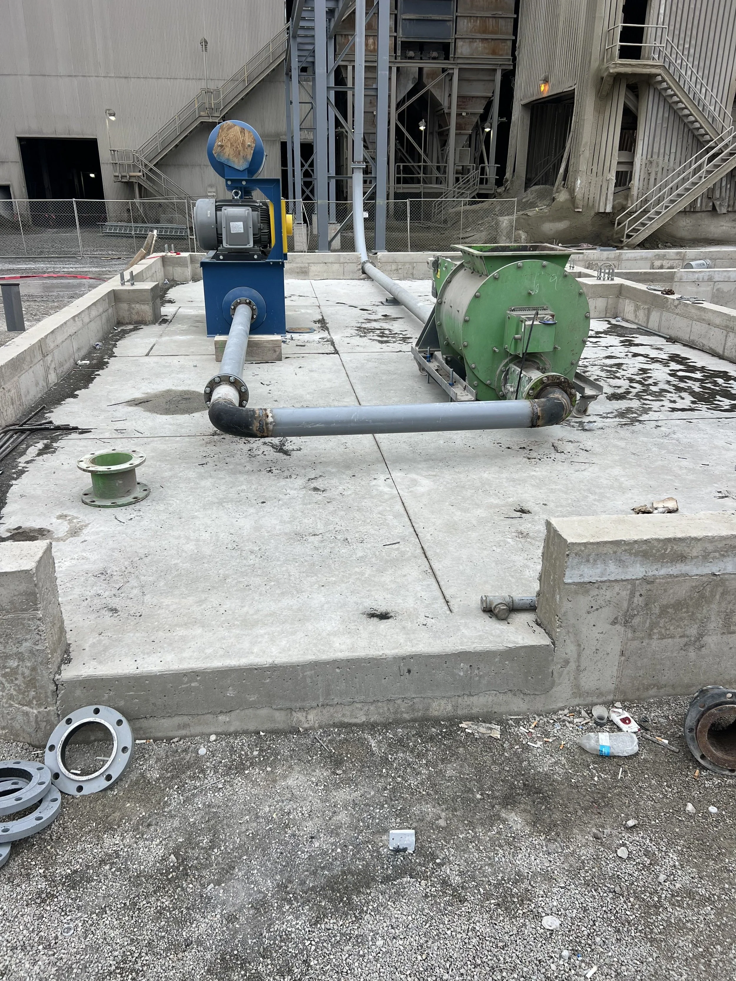 Construction site with pipes and machinery on concrete ground, partially finished building in background with stairs and fencing.