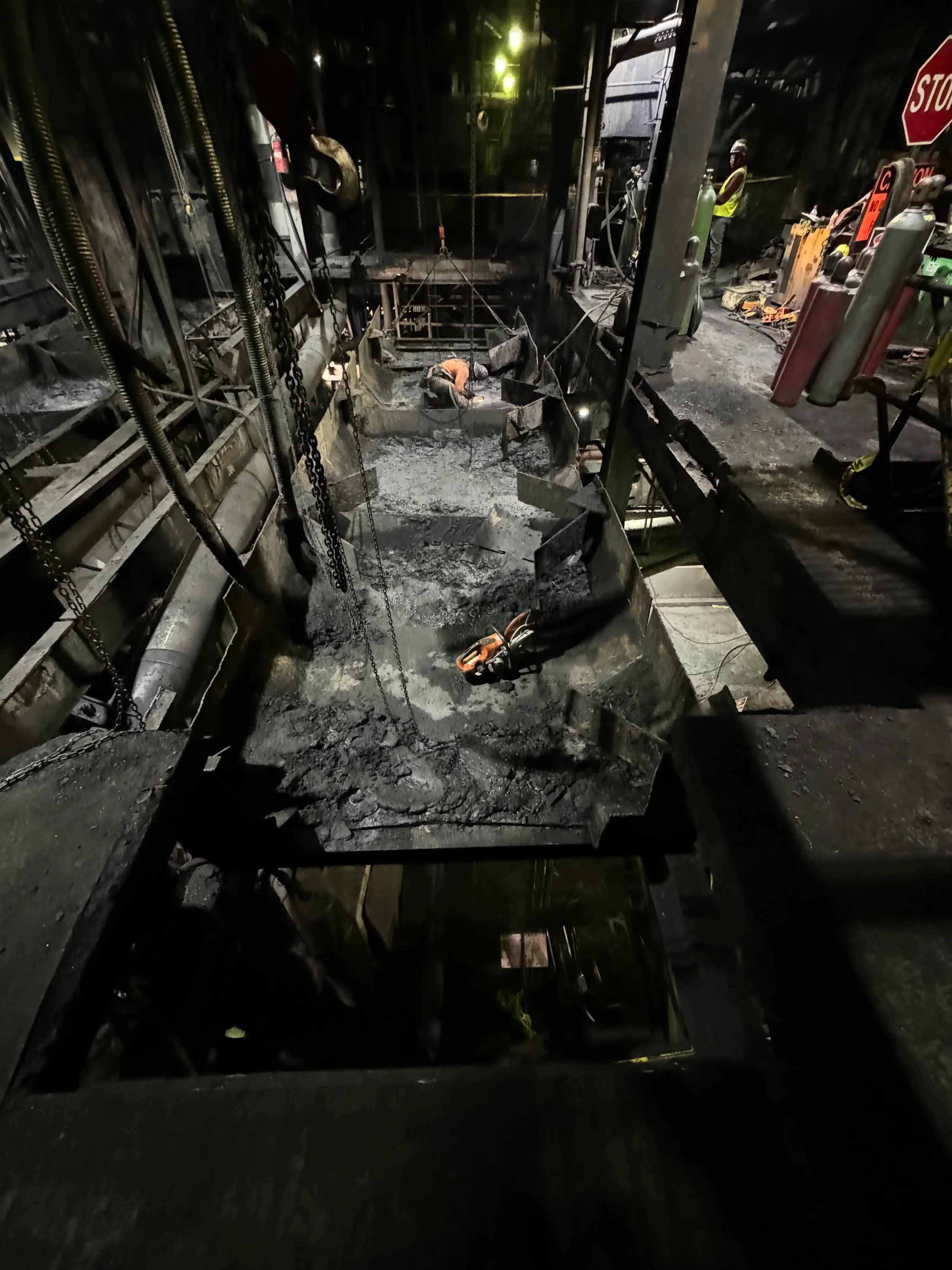 Underground construction site with workers, equipment, and debris, illuminated by yellow lighting.