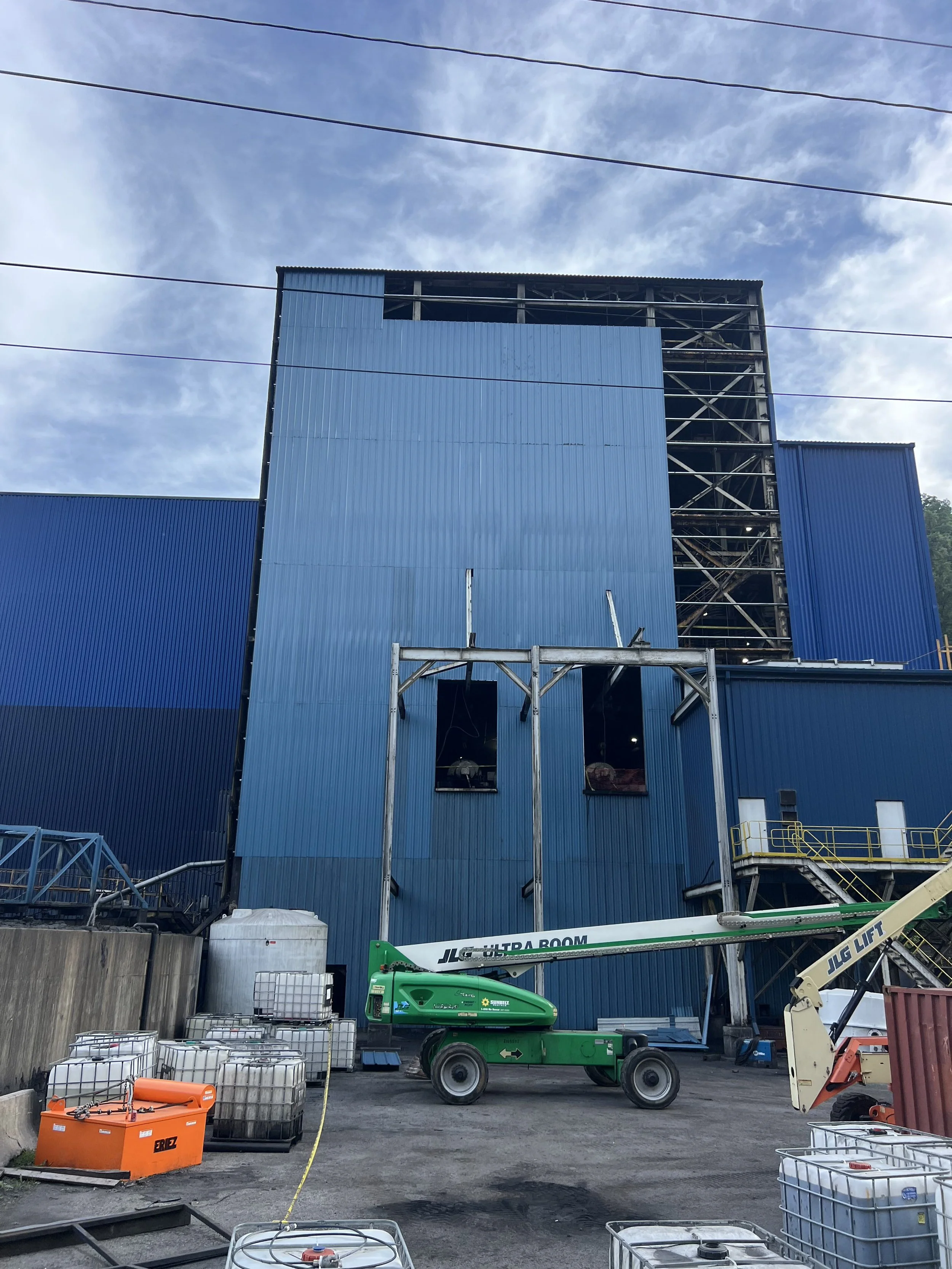 Construction site with a blue building under construction, construction equipment including a green lift, orange toolbox, and stacked water tanks in the foreground, with a partly cloudy sky overhead.