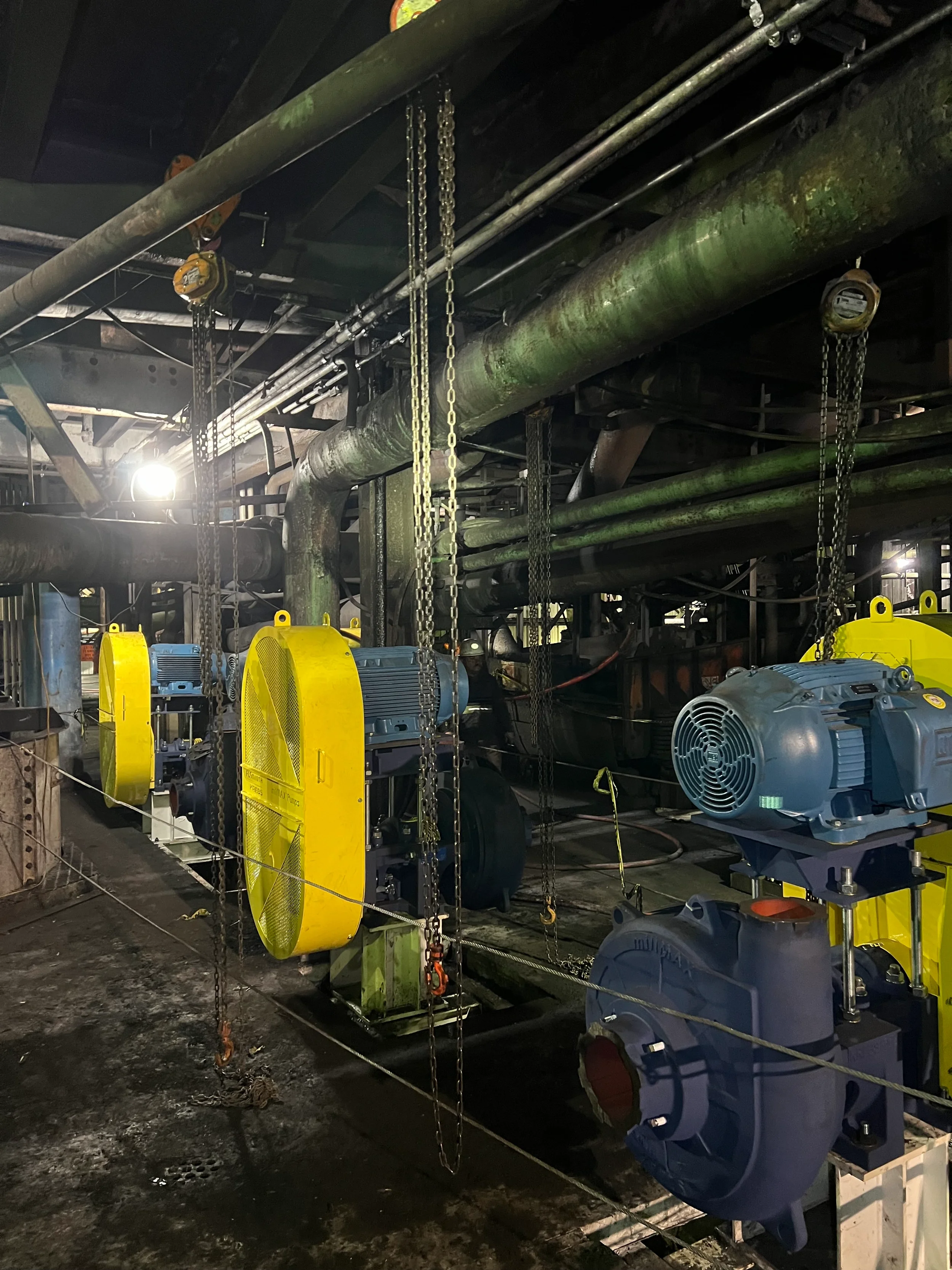 Industrial underground setting with pipes, chains, and large electric motors, featuring yellow safety guards and industrial equipment.