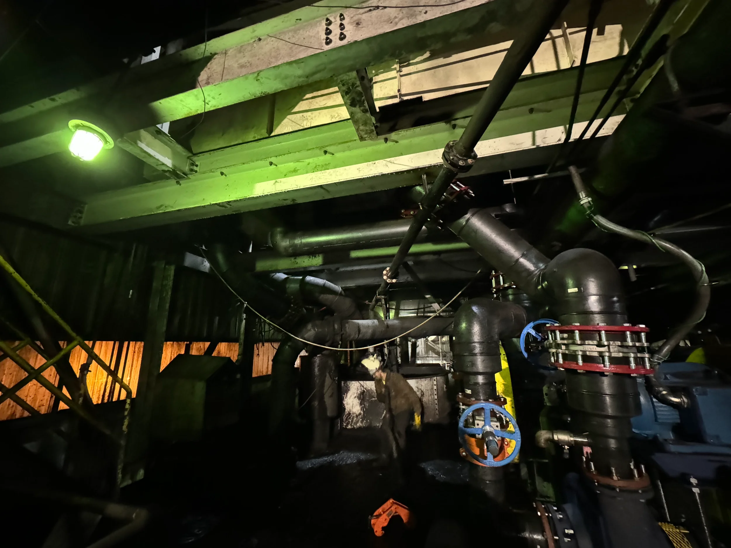 An industrial piping system with various pipes, valves, and fittings in a dimly lit environment illuminated by a green light.