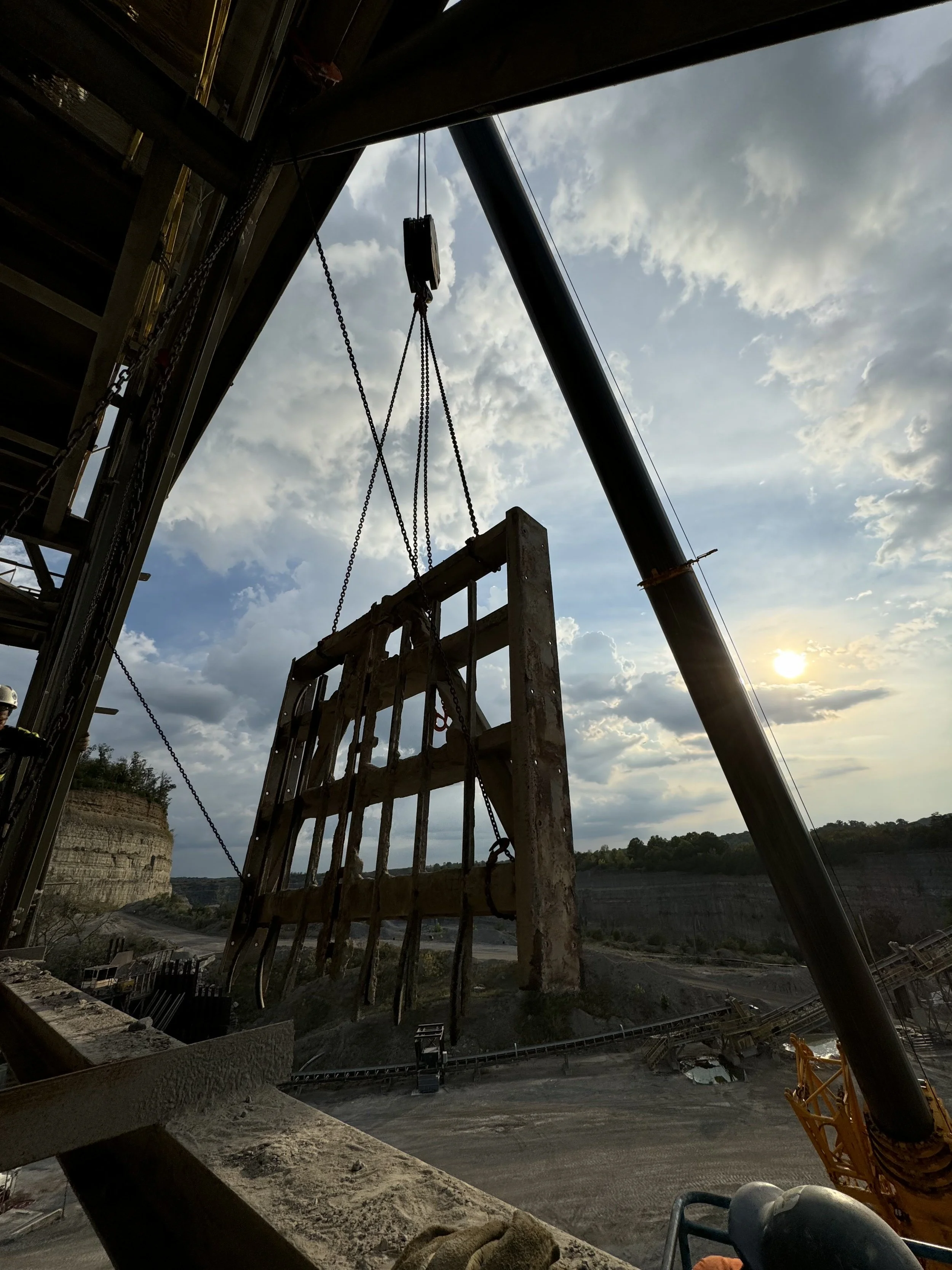 Silhouette of a construction site during sunset with steel beams and chains, overlooking a canyon and river, with cloudy sky in the background.