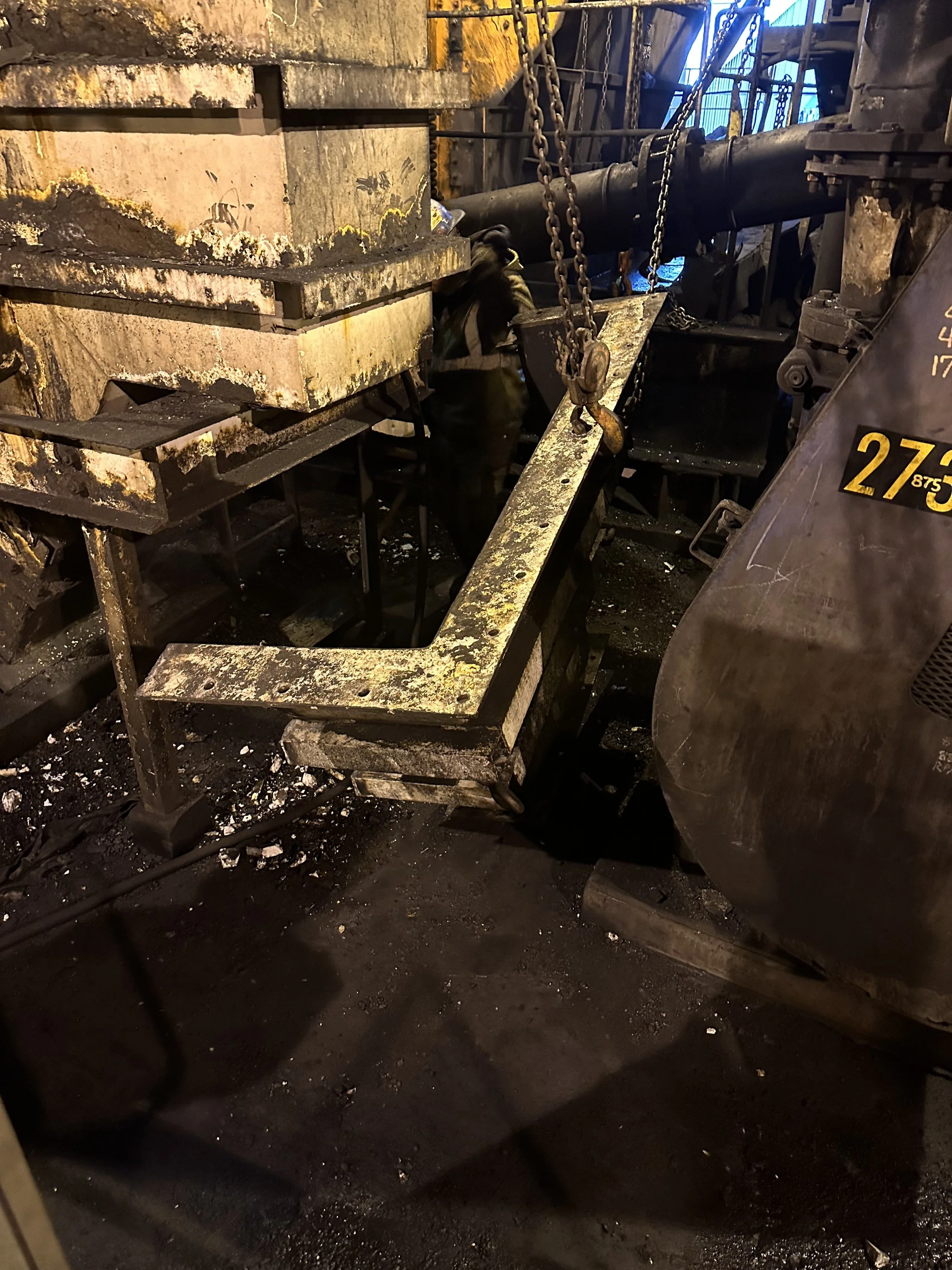 Interior of a damaged industrial facility or factory with large metal pipes, chains, and burnt debris on the floor. A person wearing black gloves is working among the machinery.