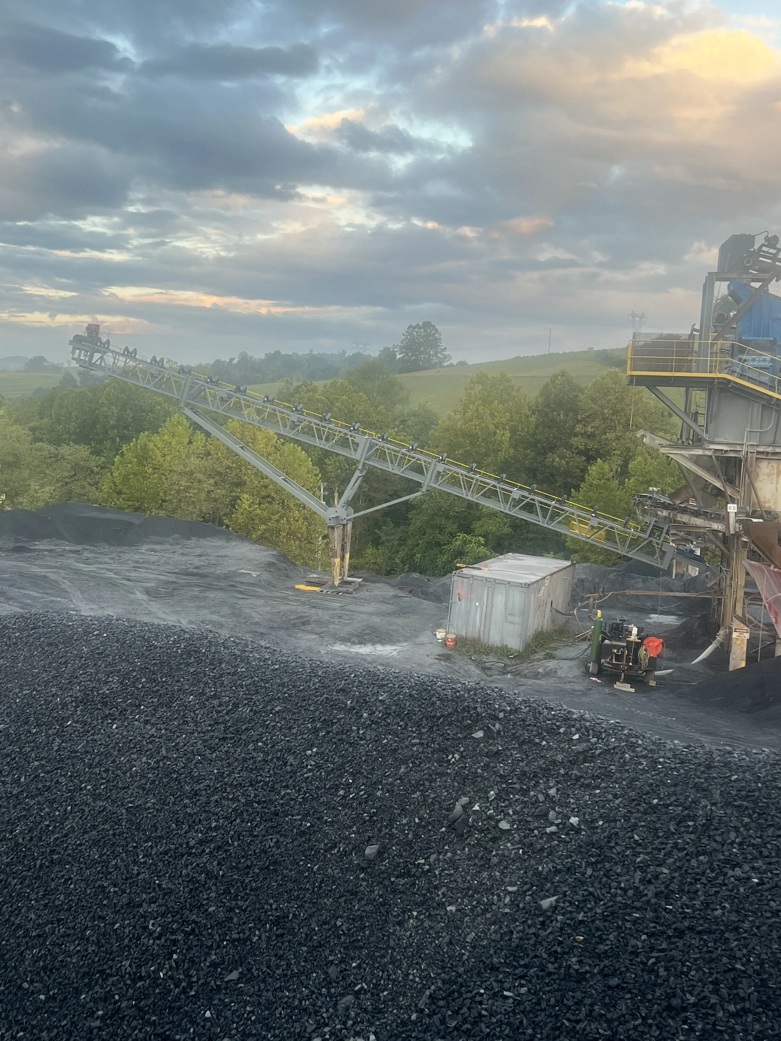 Dust pile and industrial conveyor system in a quarry with trees and hills in the background, under a partly cloudy sky at sunset.