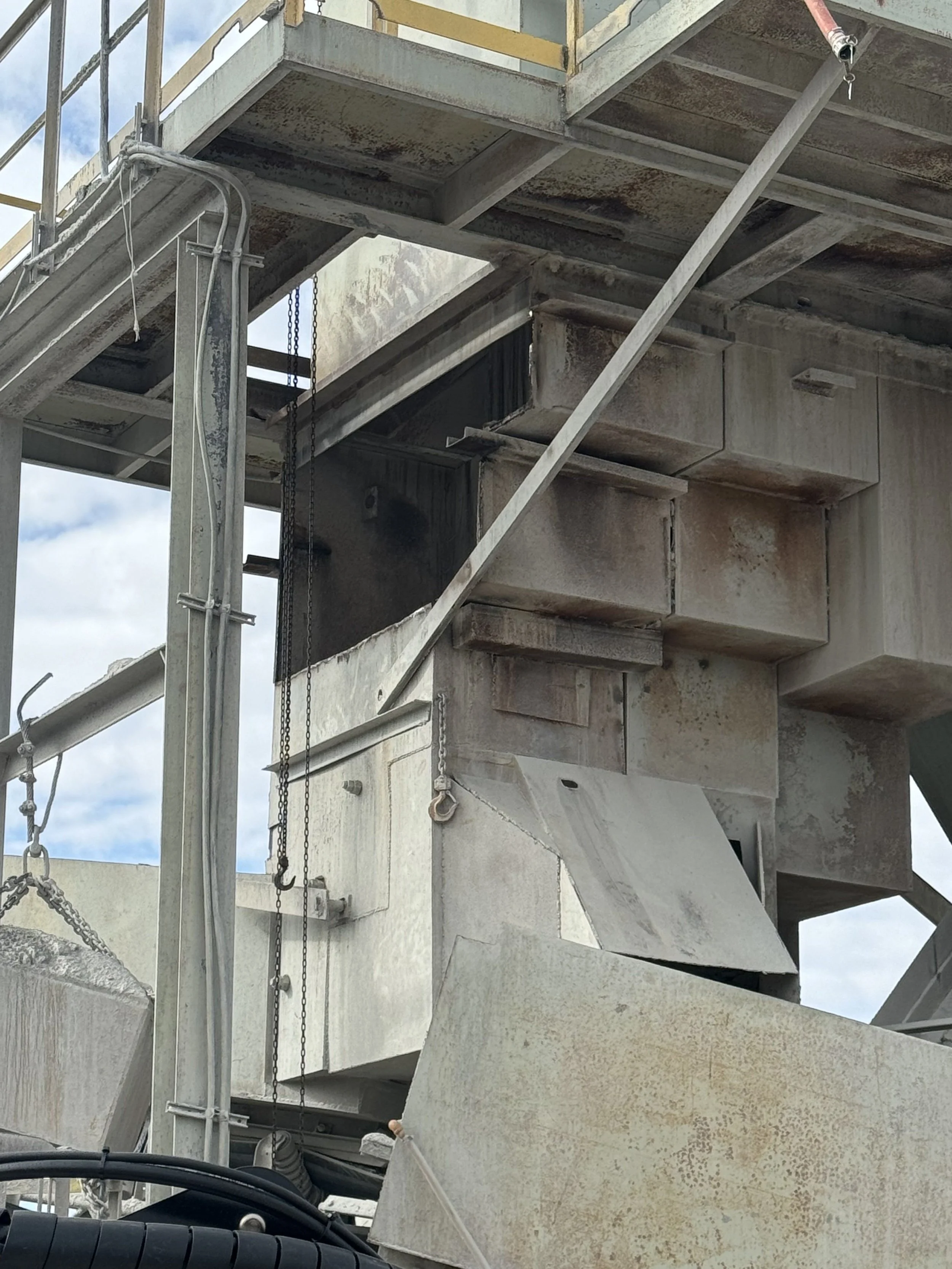 Close-up view of a construction site with large concrete and steel structures, scaffolding, and chains, under a cloudy sky.