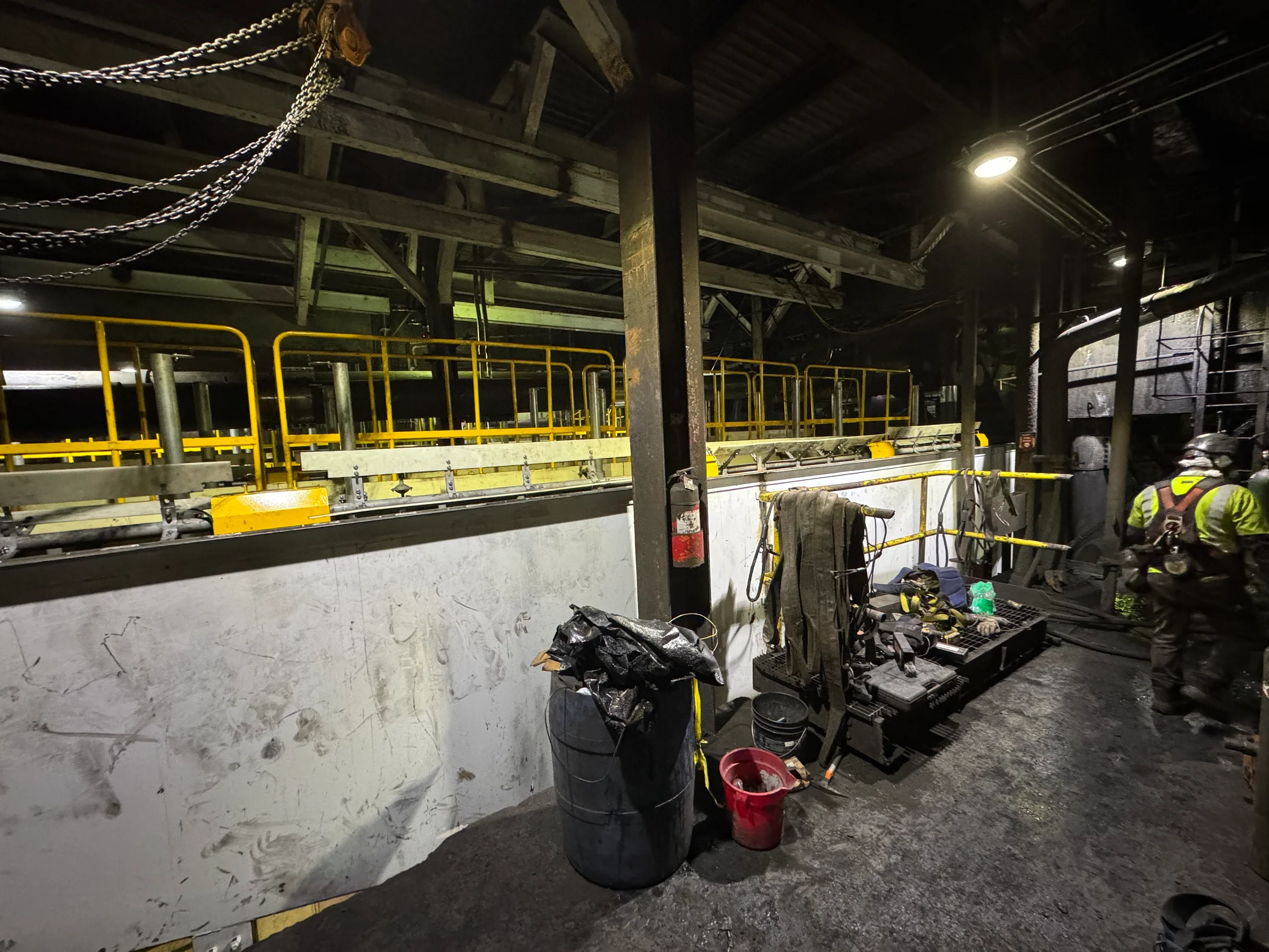 Industrial workspace with yellow safety railing, tools, and equipment. Workers in safety vests are working near a wall with pipes and chains hanging from the ceiling. The environment appears gritty and used, with industrial lighting.