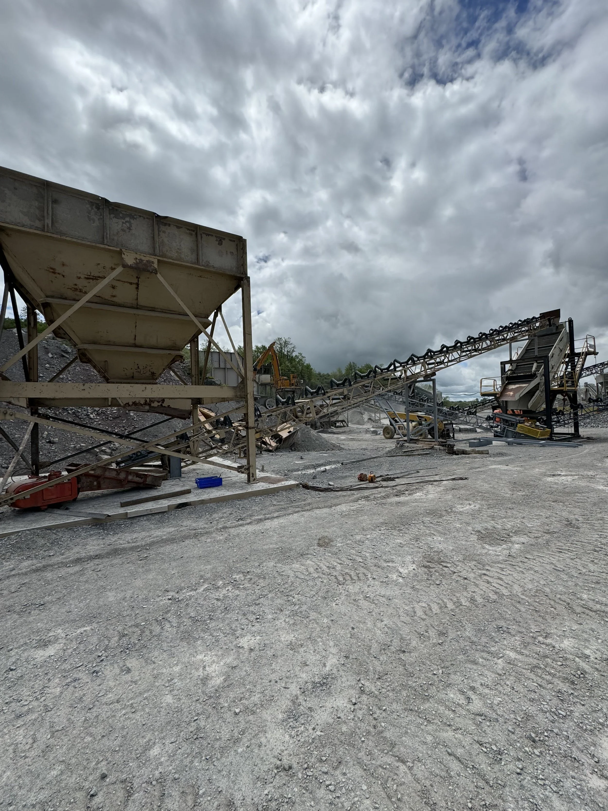 Industrial equipment and machinery in a gravelly outdoor site, with cloudy sky overhead.