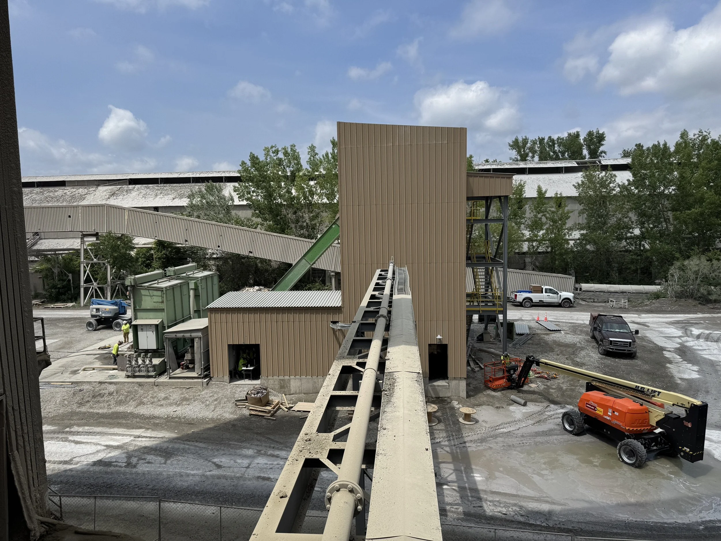 View from a construction site showing a partially built wooden structure, construction workers, and construction equipment, with a blue sky and some trees in the background.