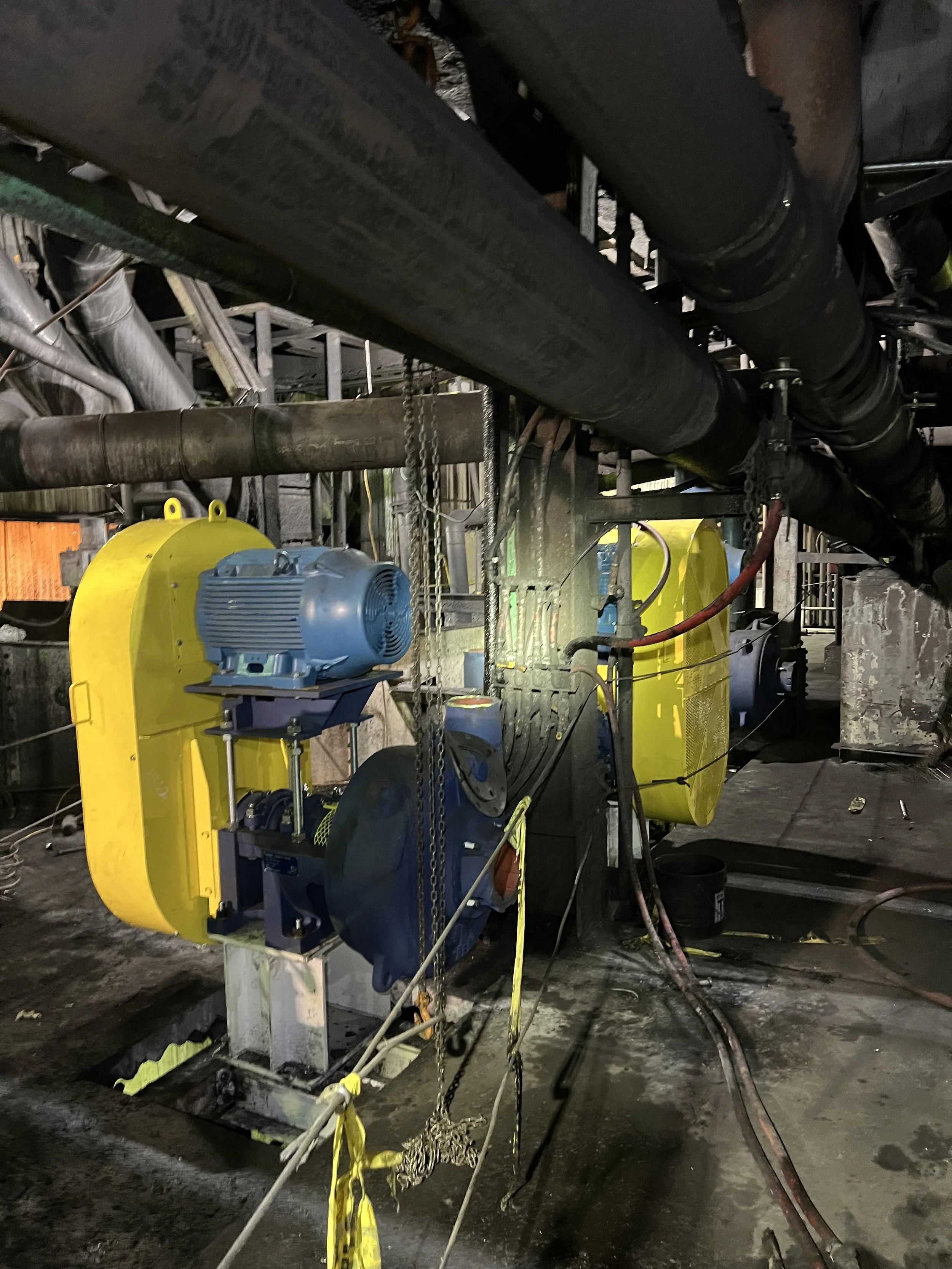 Industrial setting with large black pipes above, yellow machinery, a blue motor, chains, and cables on a concrete floor.