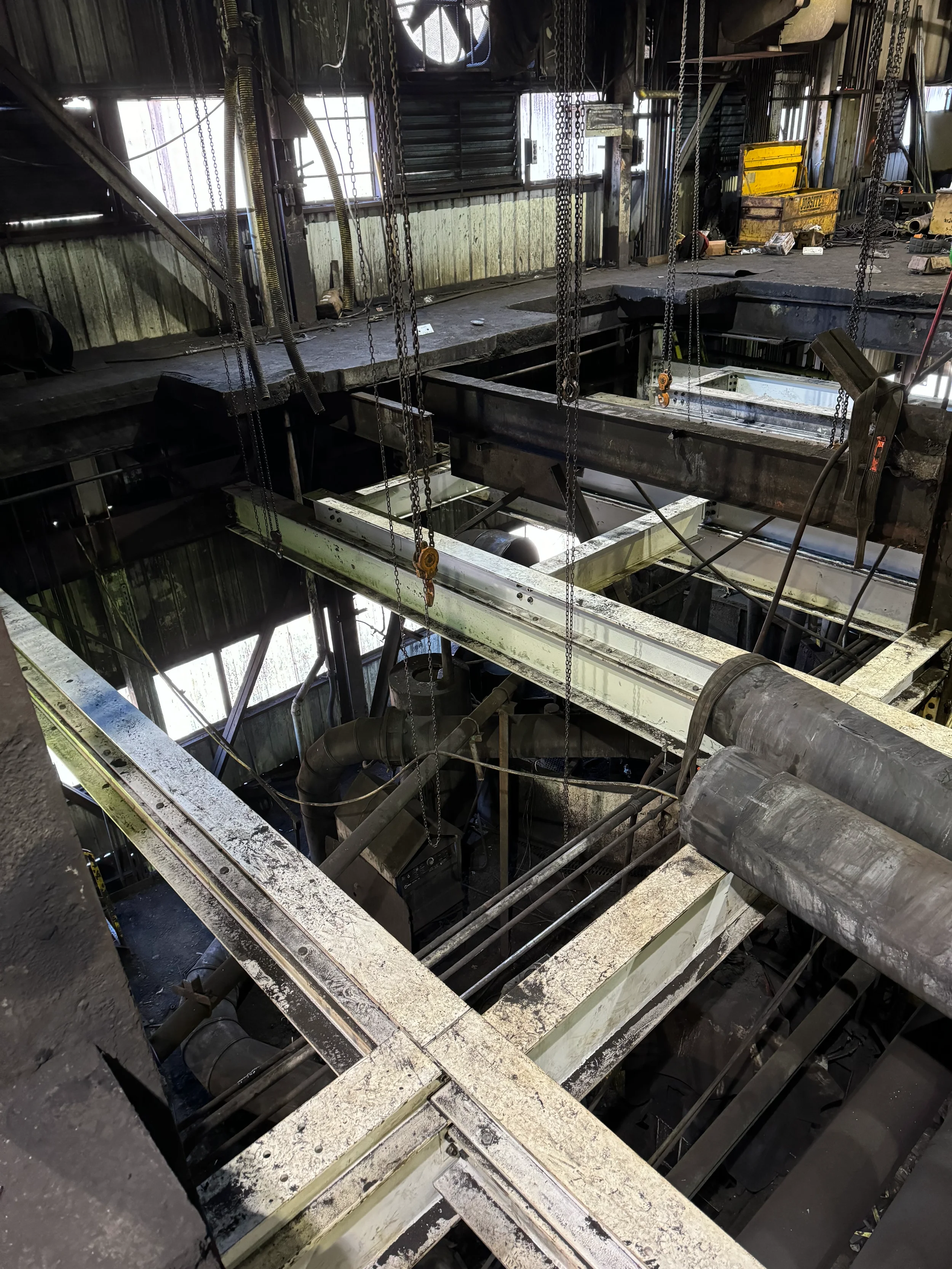View of an industrial workshop with metal beams, chains, pipes, and tools. The space appears used for metalworking or fabrication with equipment and clutter throughout.