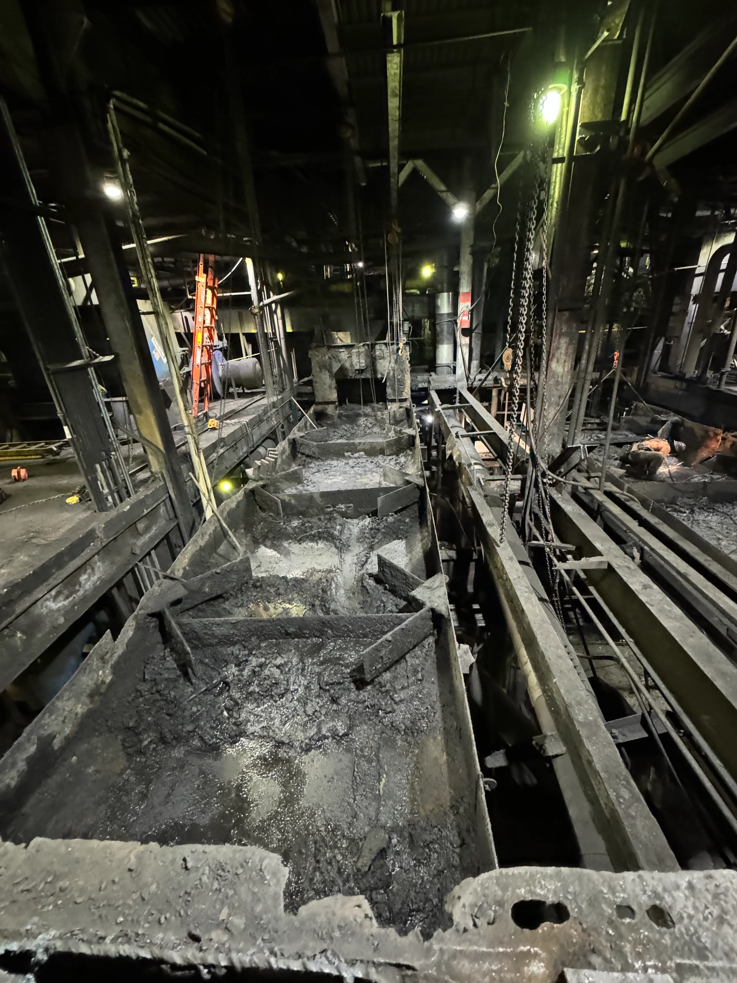 Interior view of an industrial or construction site with a conveyor belt carrying burnt or charred materials, metal scaffolding, chains, and illuminated equipment, all covered in soot and ash.