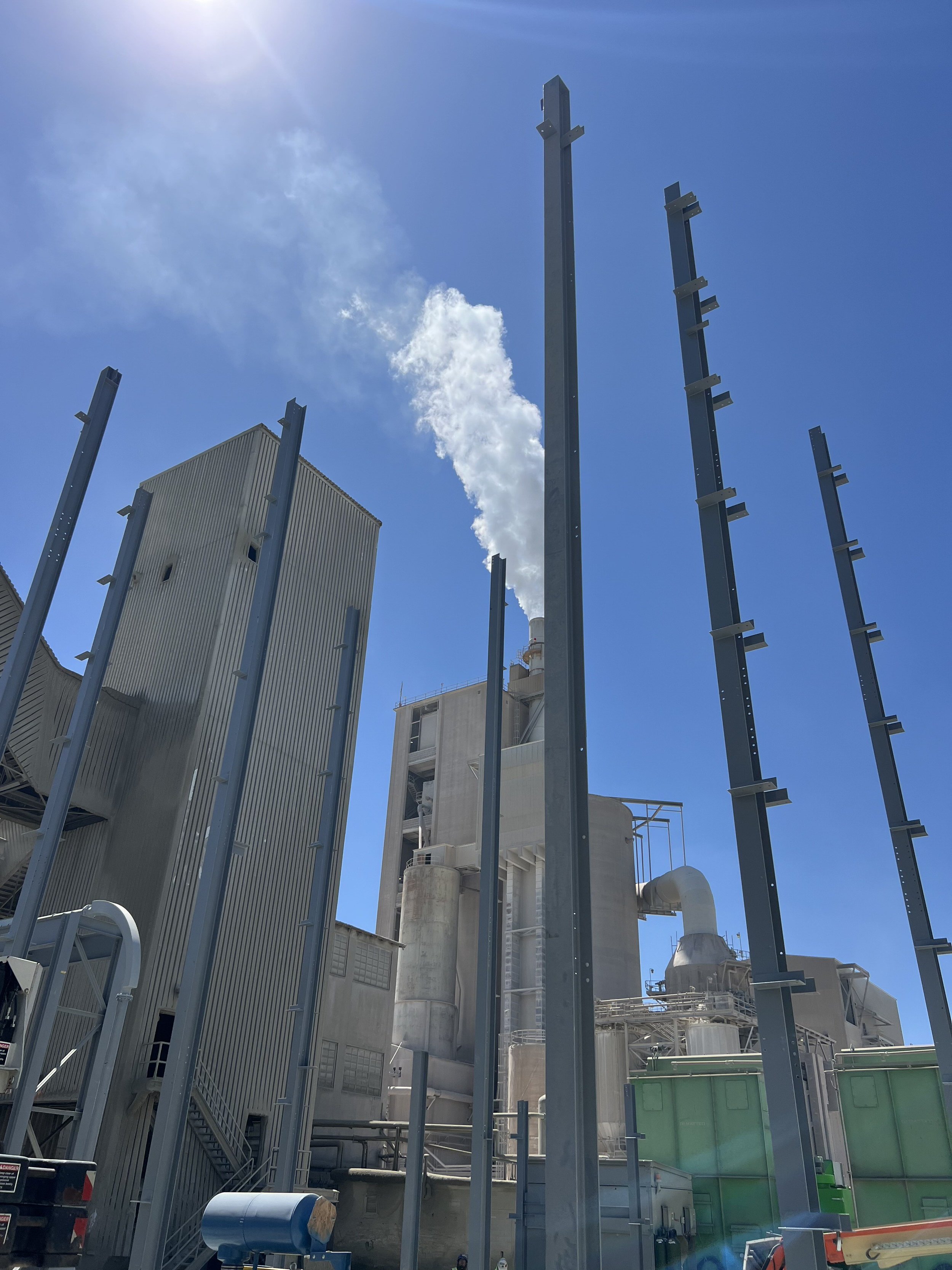 Industrial facility with tall metal structures, pipes, and a building with a smokestack emitting white smoke, under a blue sky with some clouds.