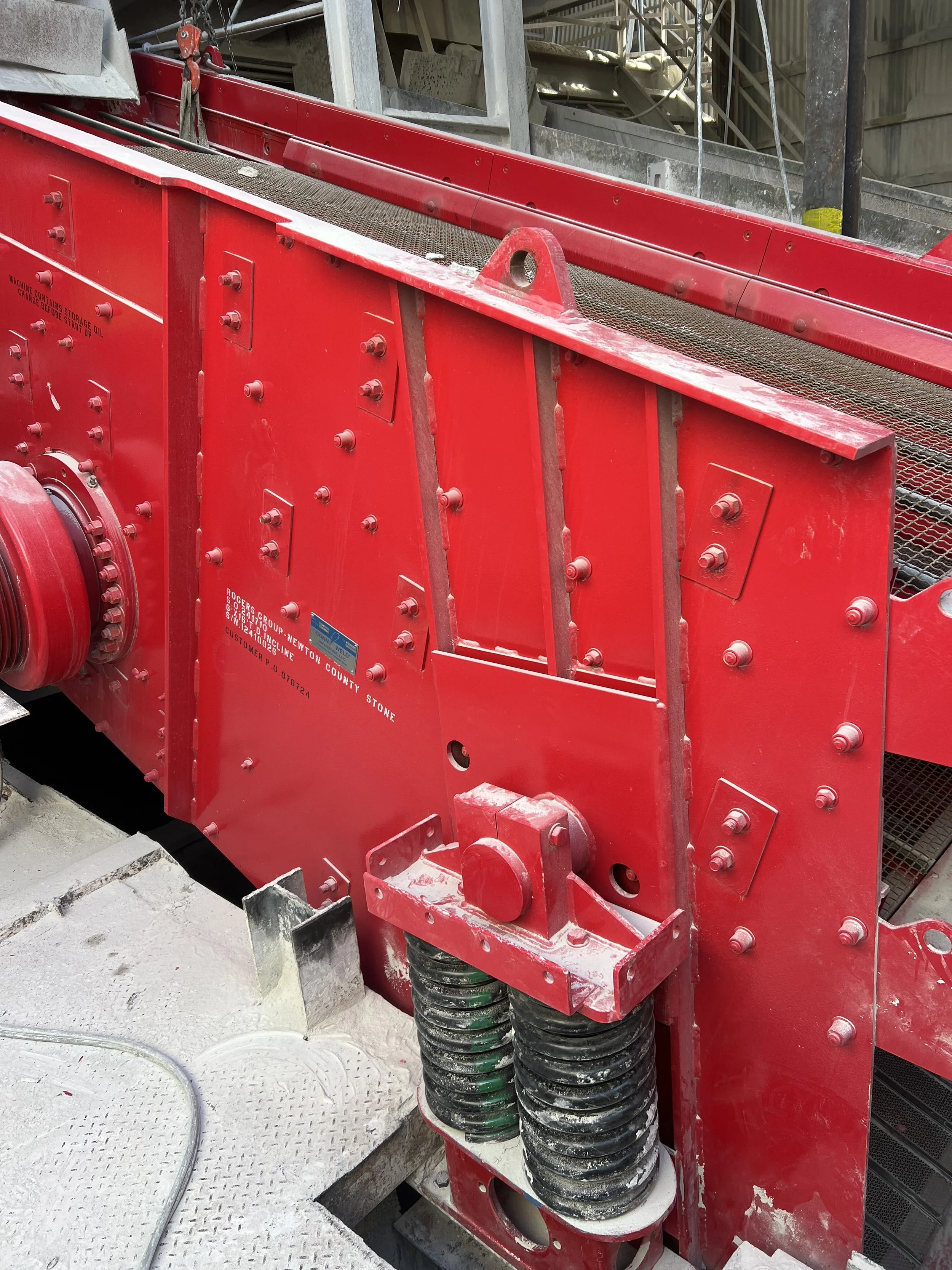 Close-up of red industrial machinery with black coiled hoses, metallic bolts, and a mesh conveyor belt in the background, located in an industrial setting.