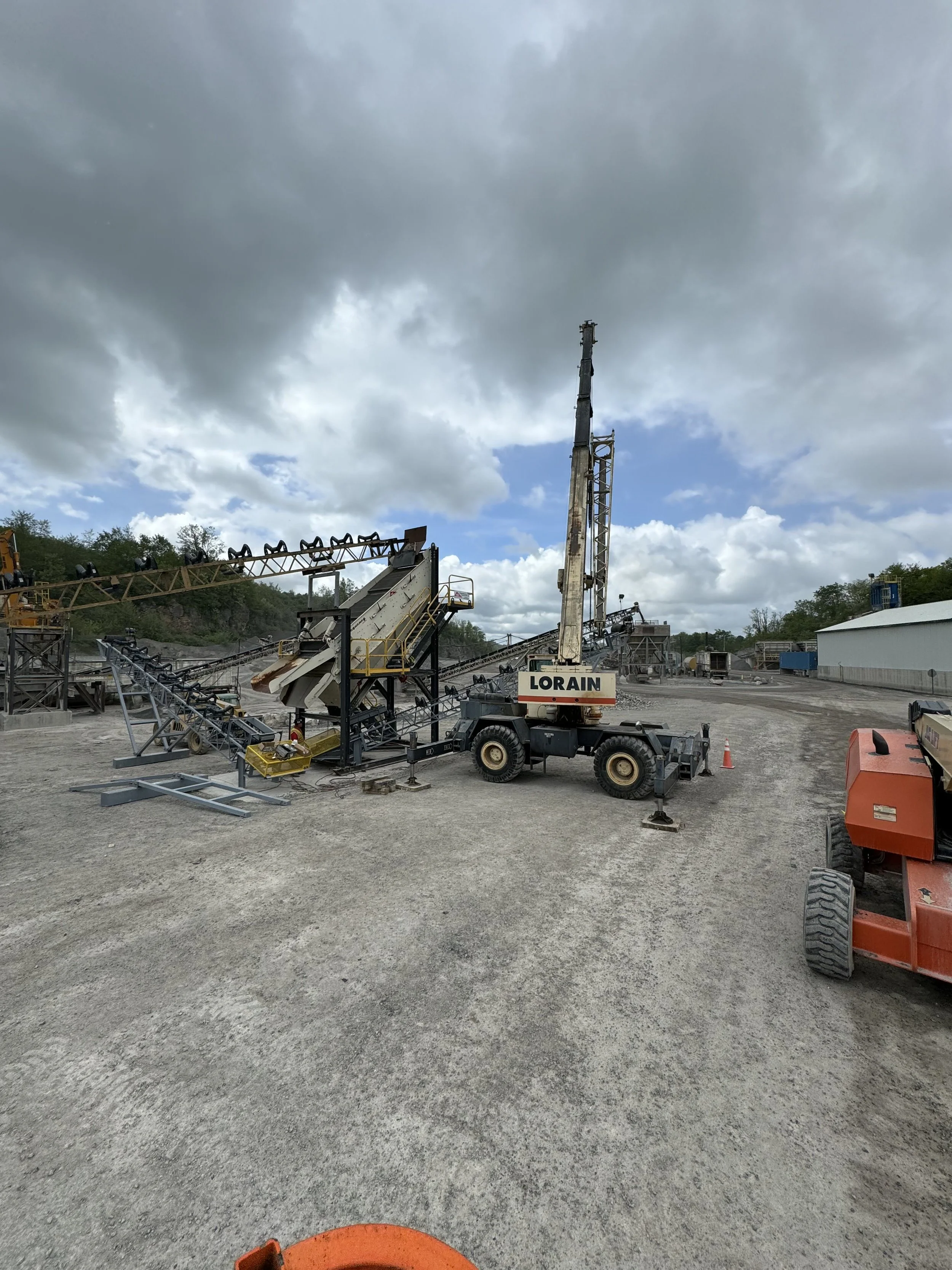 Construction site with heavy machinery, including a tall crane, conveyor belts, and equipment against a cloudy sky.