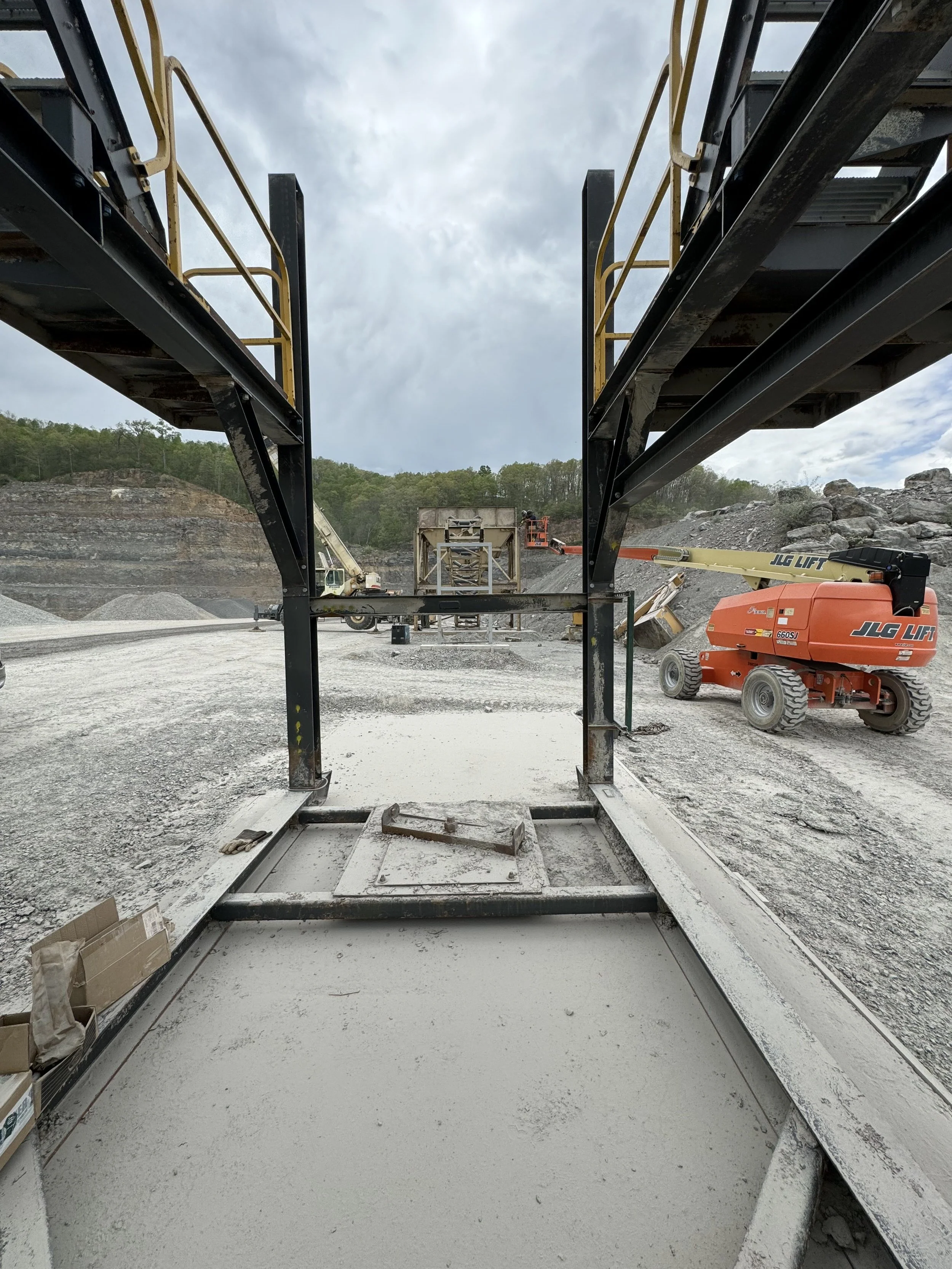 A construction site with two large conveyor belts facing each other, with a gravel or dirt surface underneath. In the background, there are hills covered with trees, and the sky is cloudy. There is construction machinery, including a JLG lift and ano