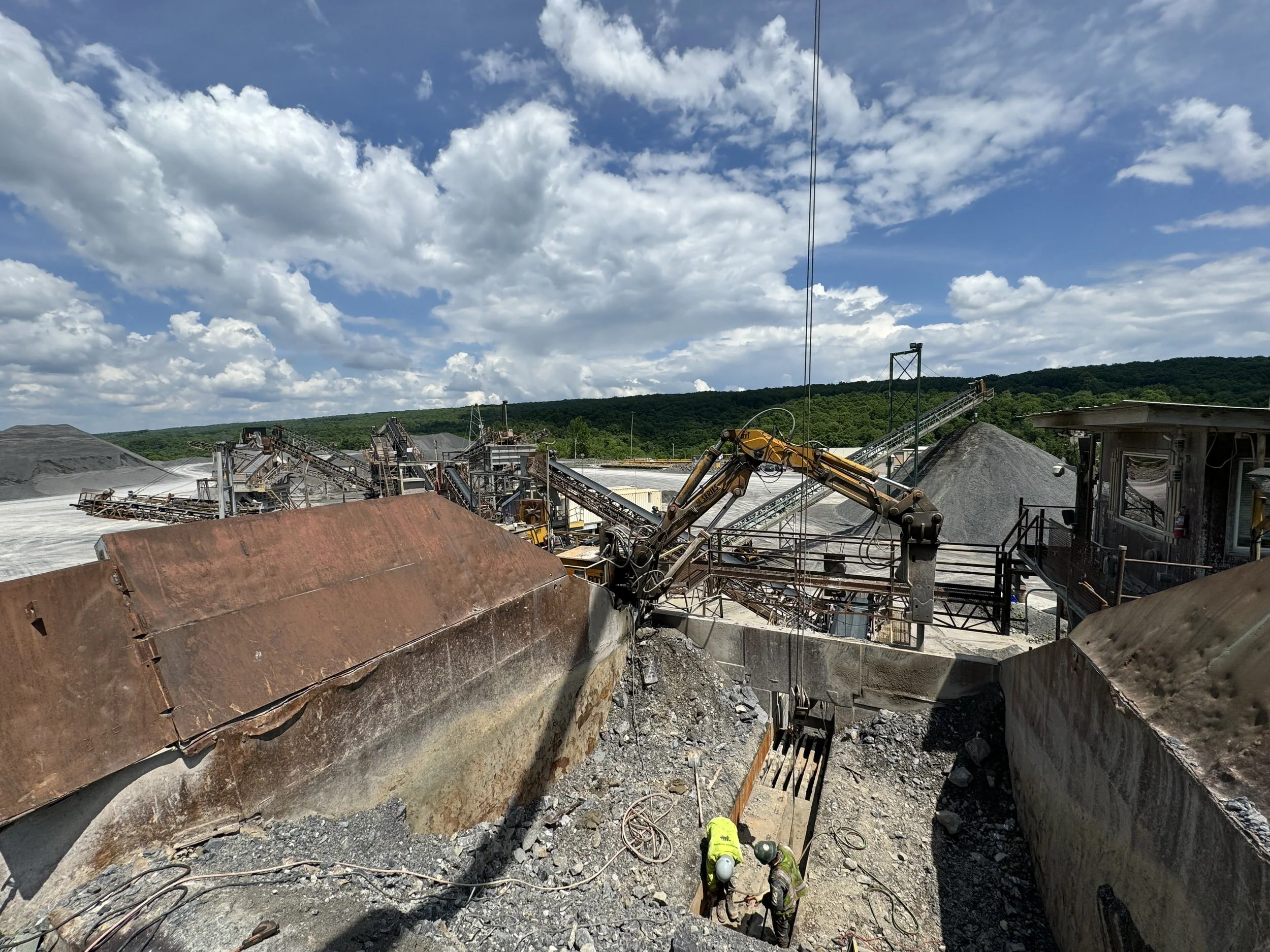 Construction site with machinery and workers, large rocks, and gravel piles, with a background of greenery and a partly cloudy sky.