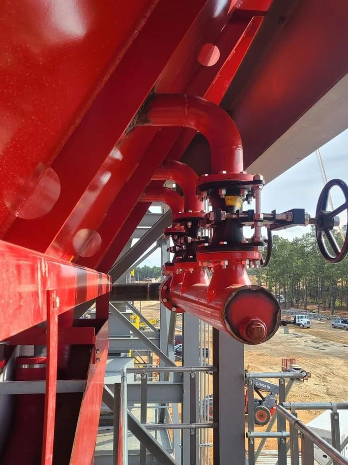 Close-up of red industrial pipes and valves on a construction site with a view of an open area with vehicles and trees in the background.