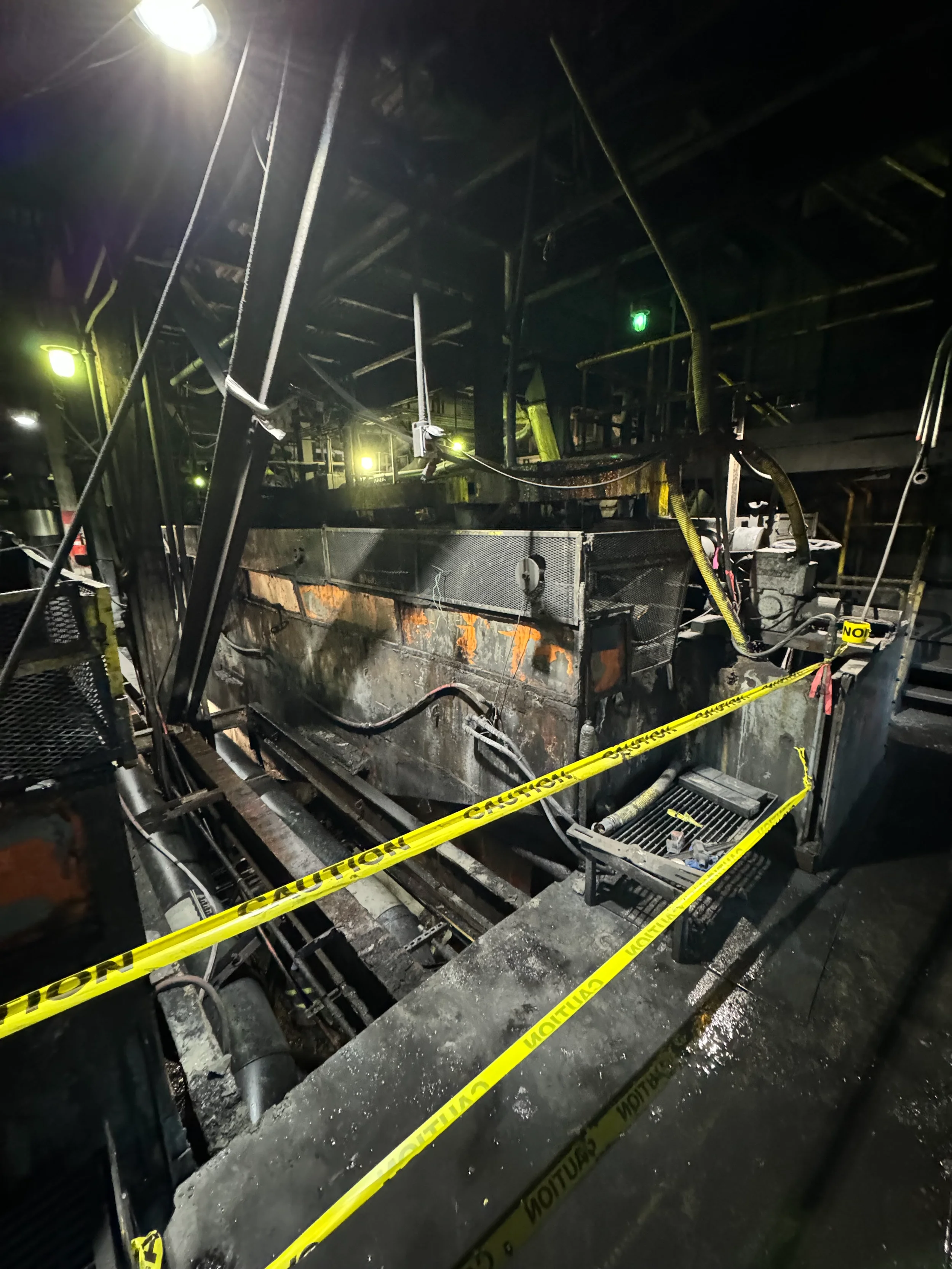 Burned industrial machine or vehicle with charred metal, surrounded by toxic caution tape in a dark environment.