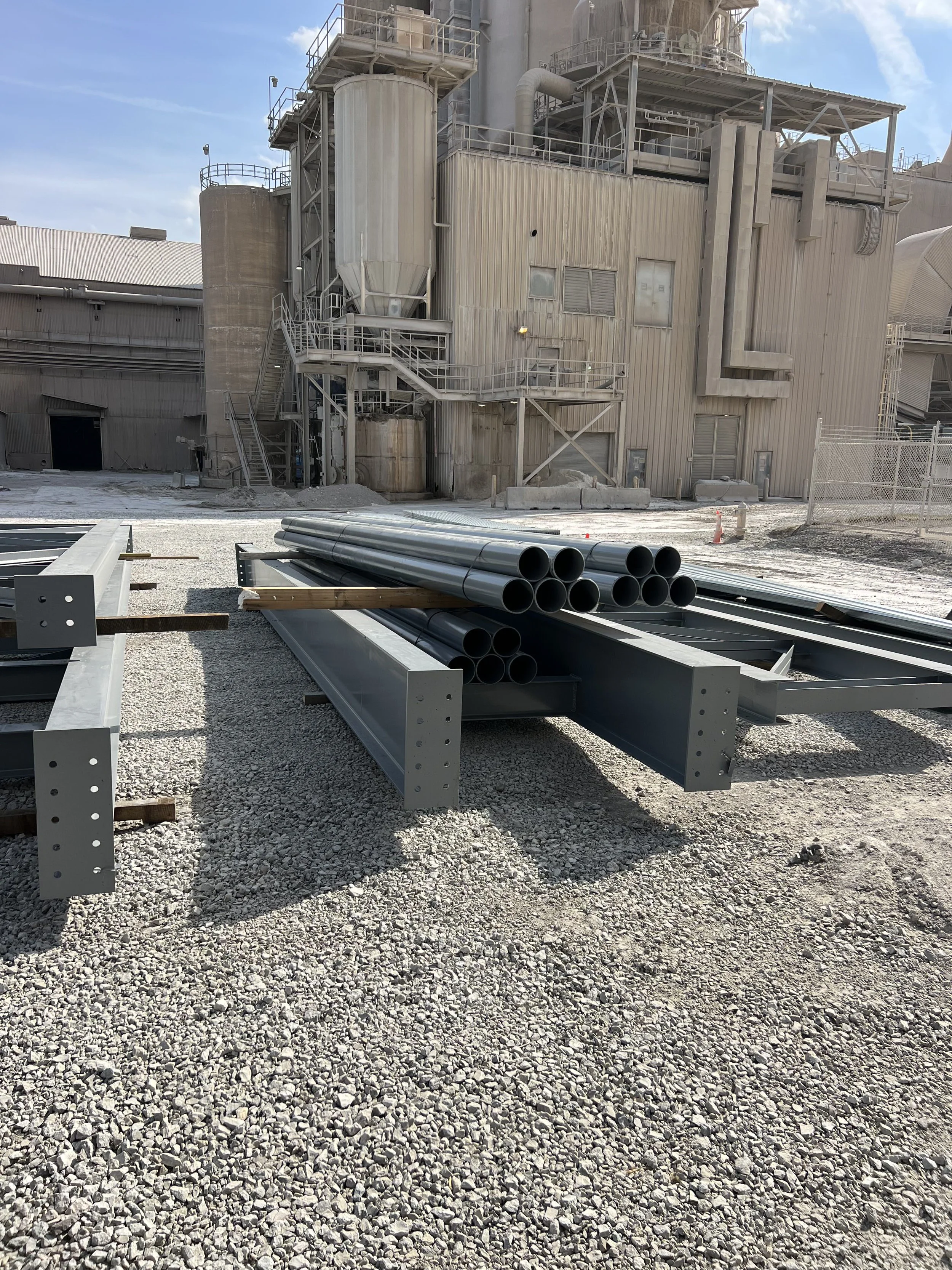 Construction site with metal pipes, steel beams, and a large industrial building in the background. Gravel ground with some construction materials and cones.