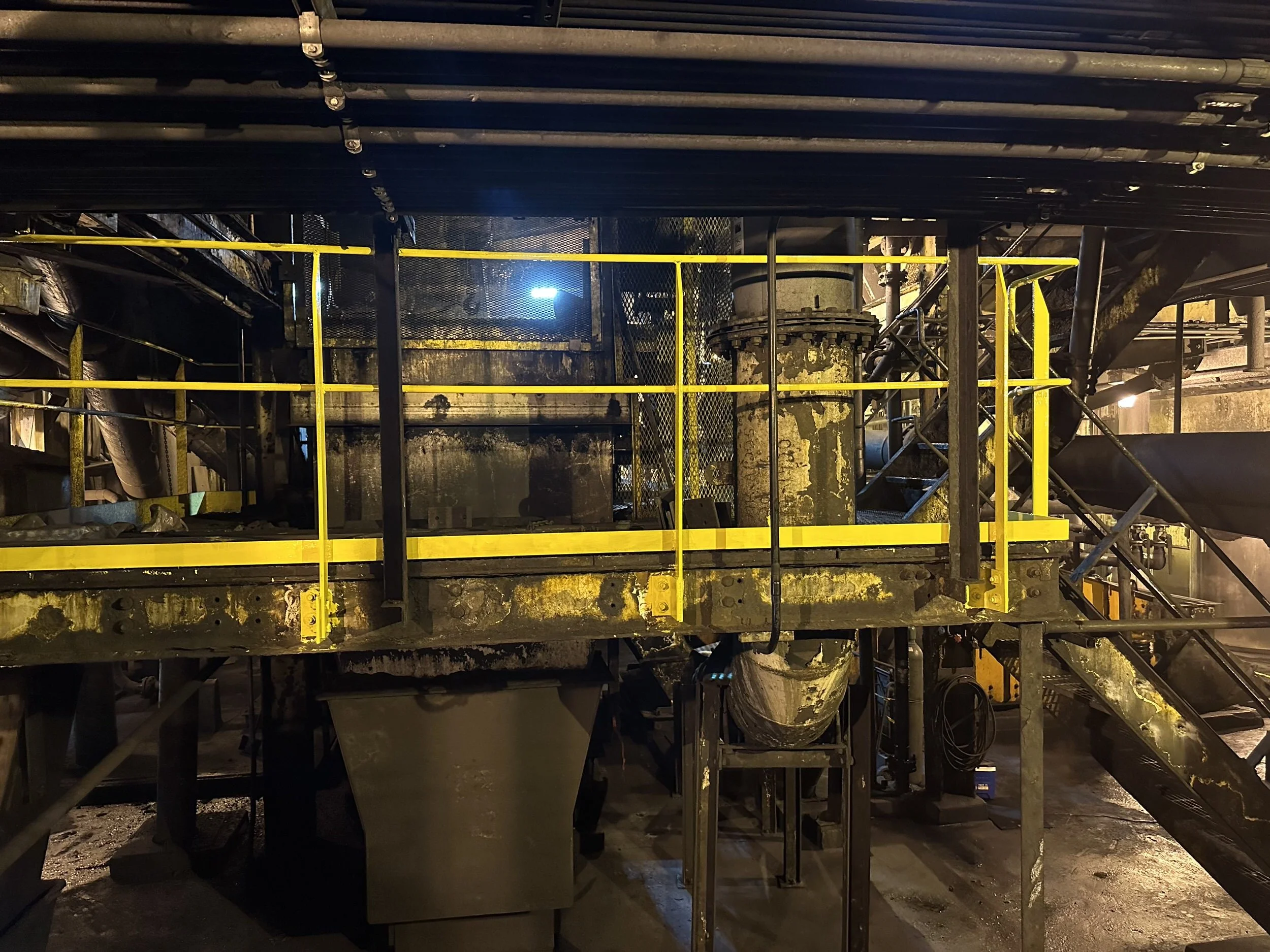 Industrial setting with metal stairs, railings, pipes, and machinery, illuminated by yellowish lighting.