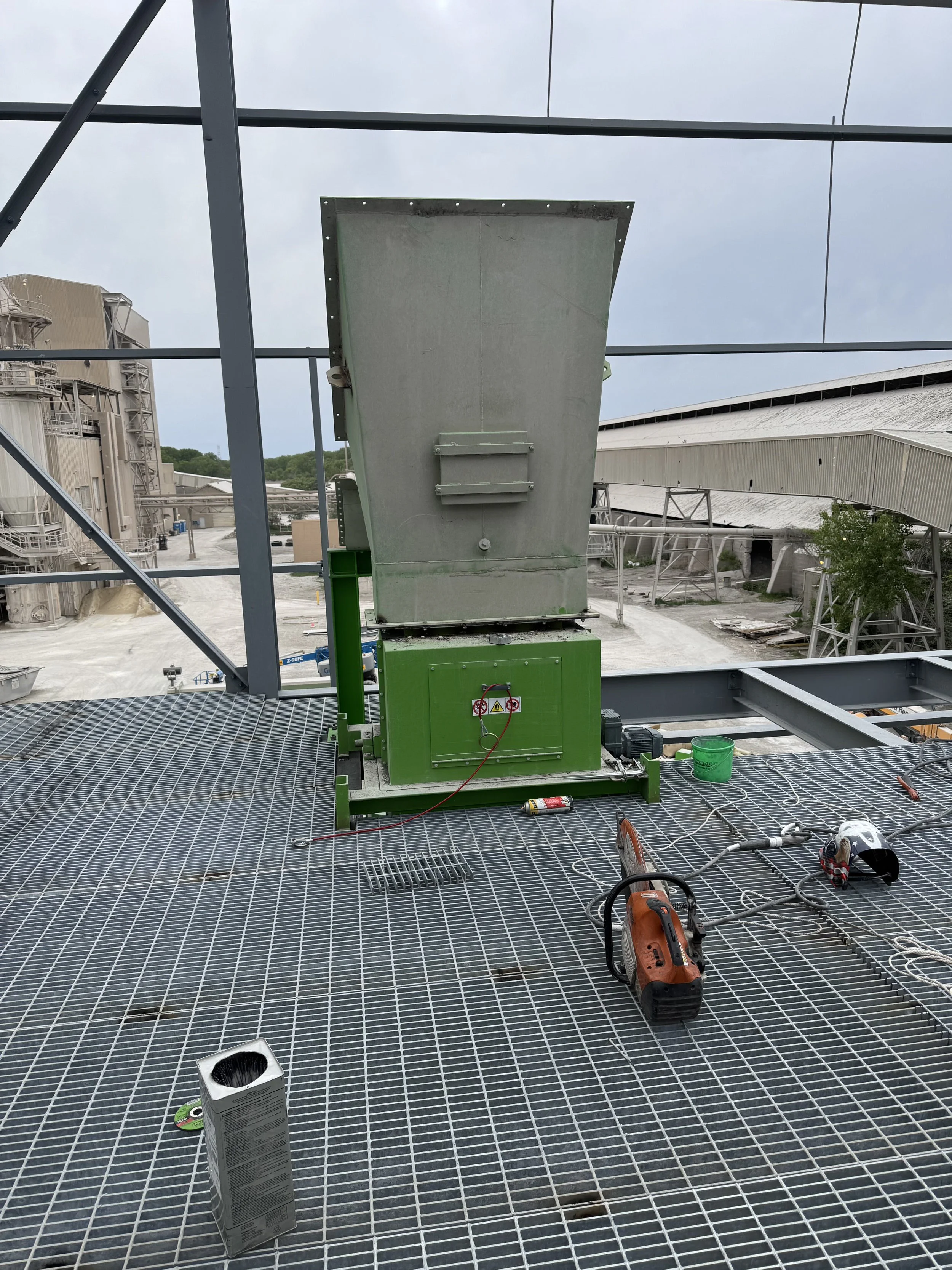 Construction site with metal grating flooring and equipment, including a green machine with a large tilted metal surface, tools, and safety gear, with industrial buildings and cloudy sky in the background.