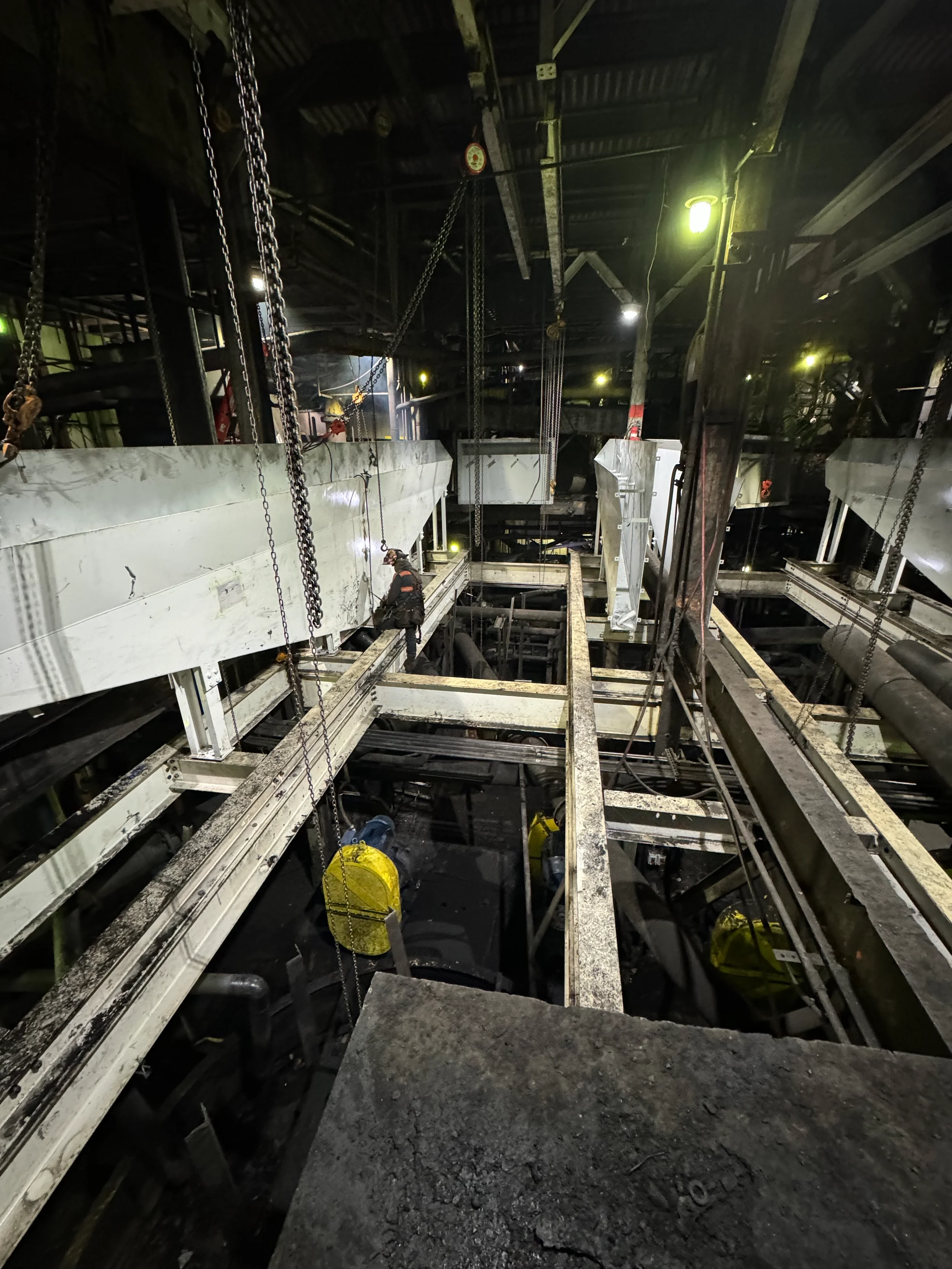 Inside a dark, industrial construction or maintenance site with various metal structures, chains, and yellow safety equipment, and a worker in safety gear.