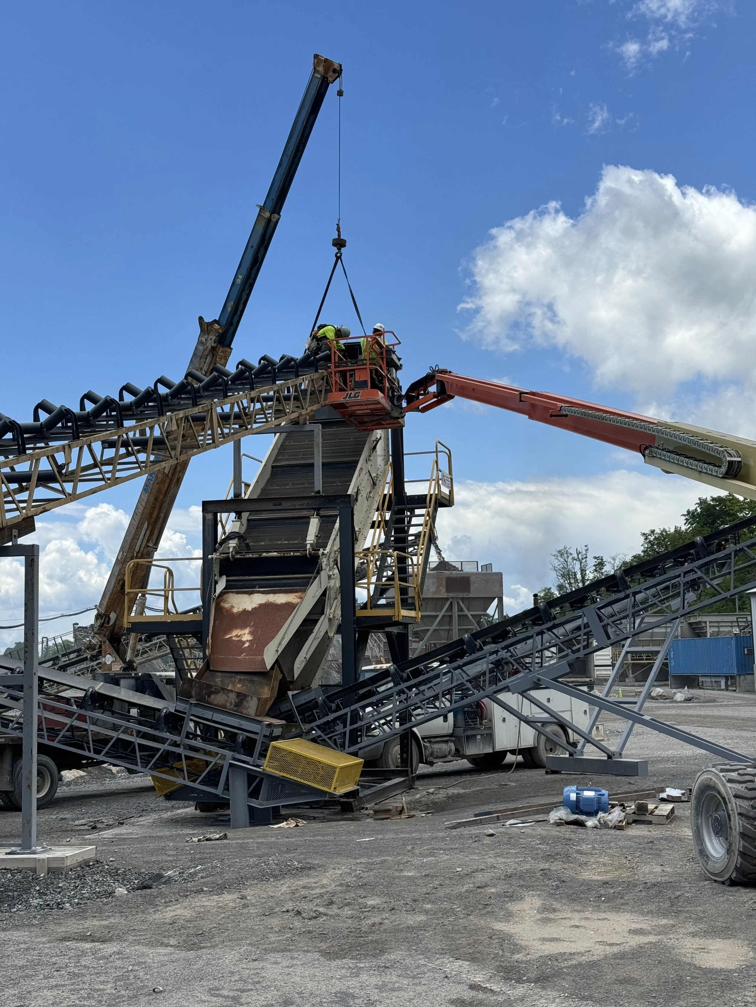 Construction site with a crane lifting equipment, workers in safety gear, conveyor belts, and machinery against a partly cloudy sky.