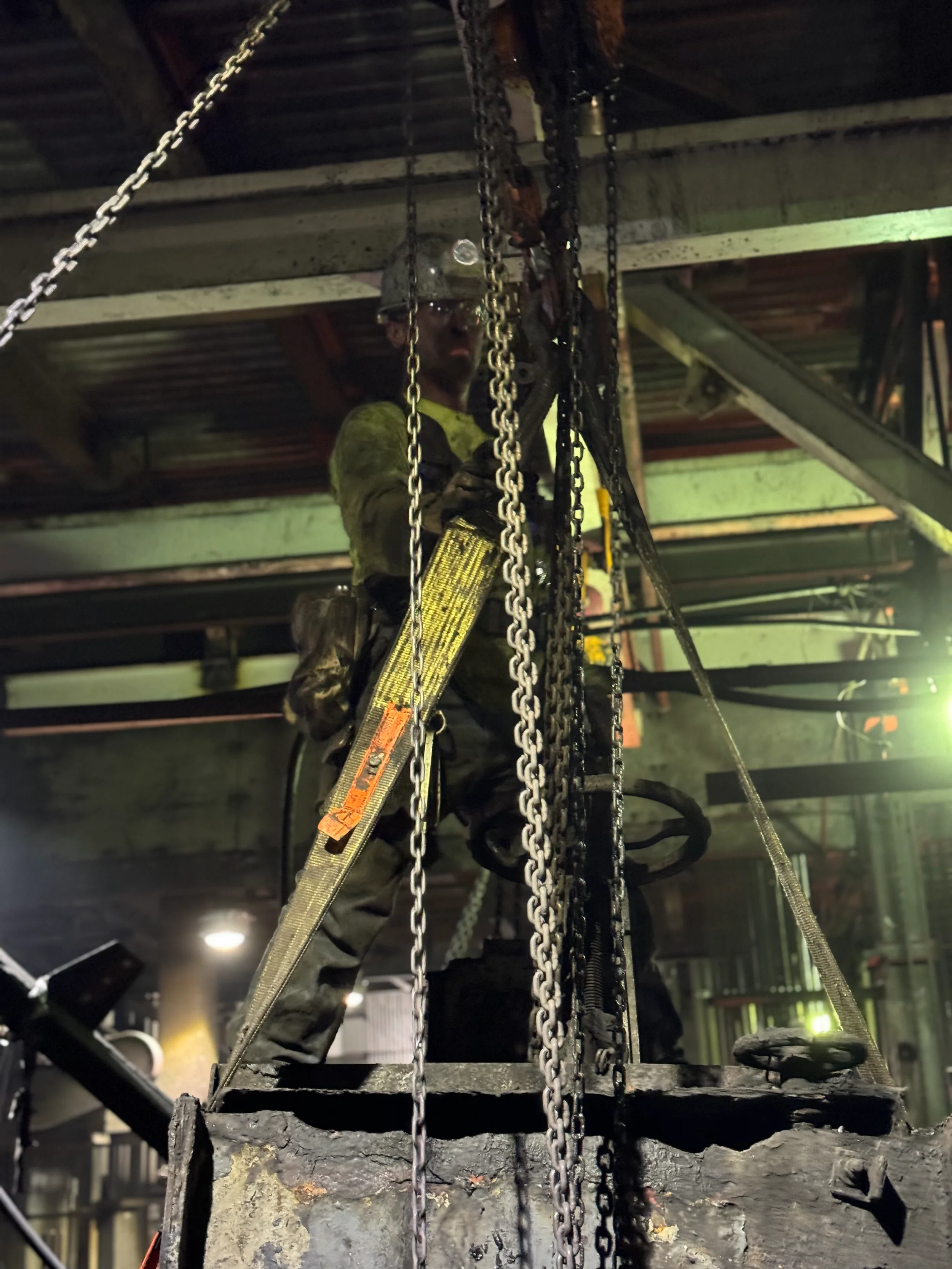 A construction worker wearing a helmet and safety gear is working on a metal structure with chains and a ladder inside an industrial or warehouse setting.