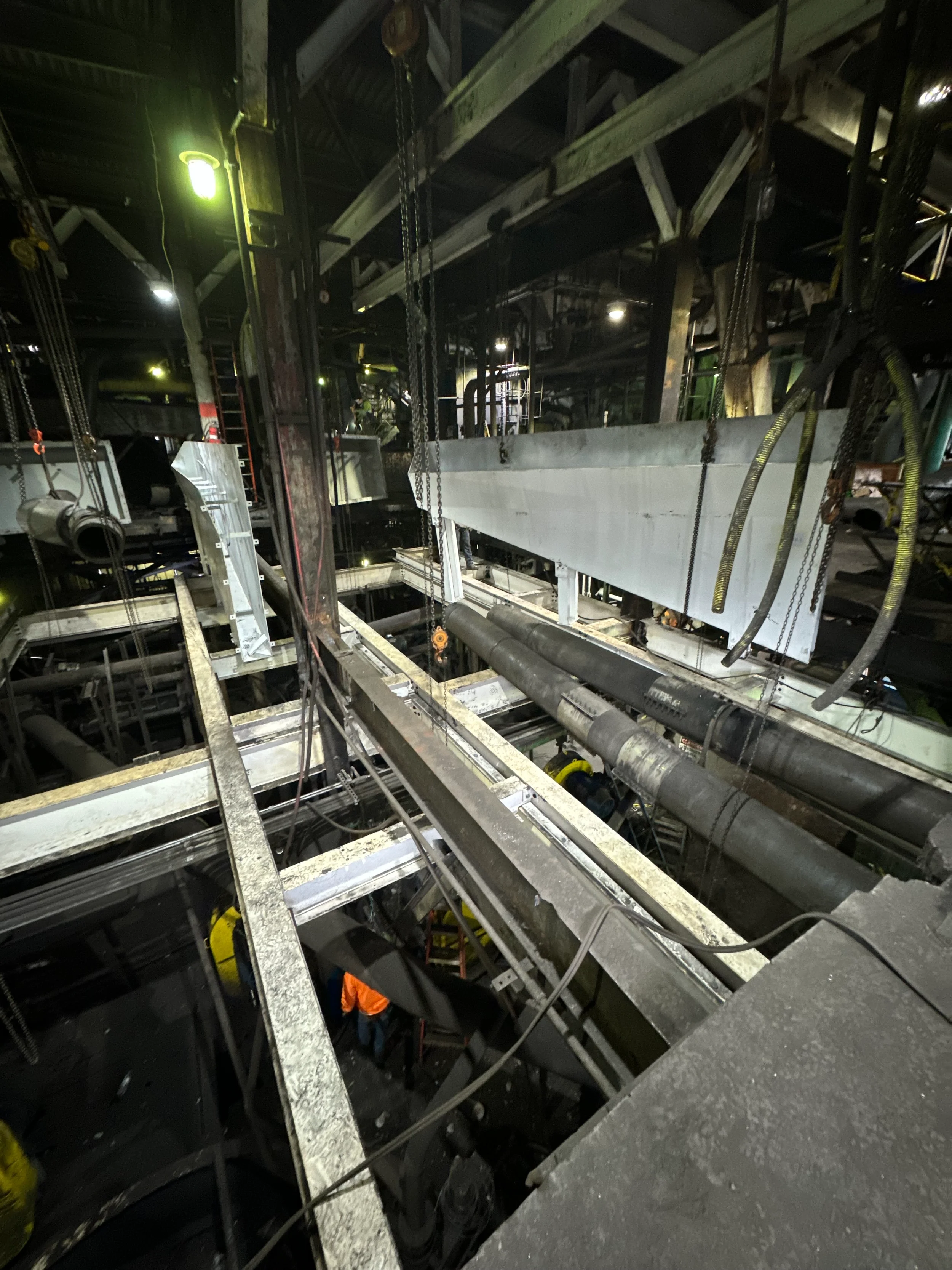 An industrial construction site in a building under construction, with metal beams, pipes, and scaffolding visible. There are chains and equipment hanging, with workers in orange safety vests below. The setting is dark, with some artificial lighting.