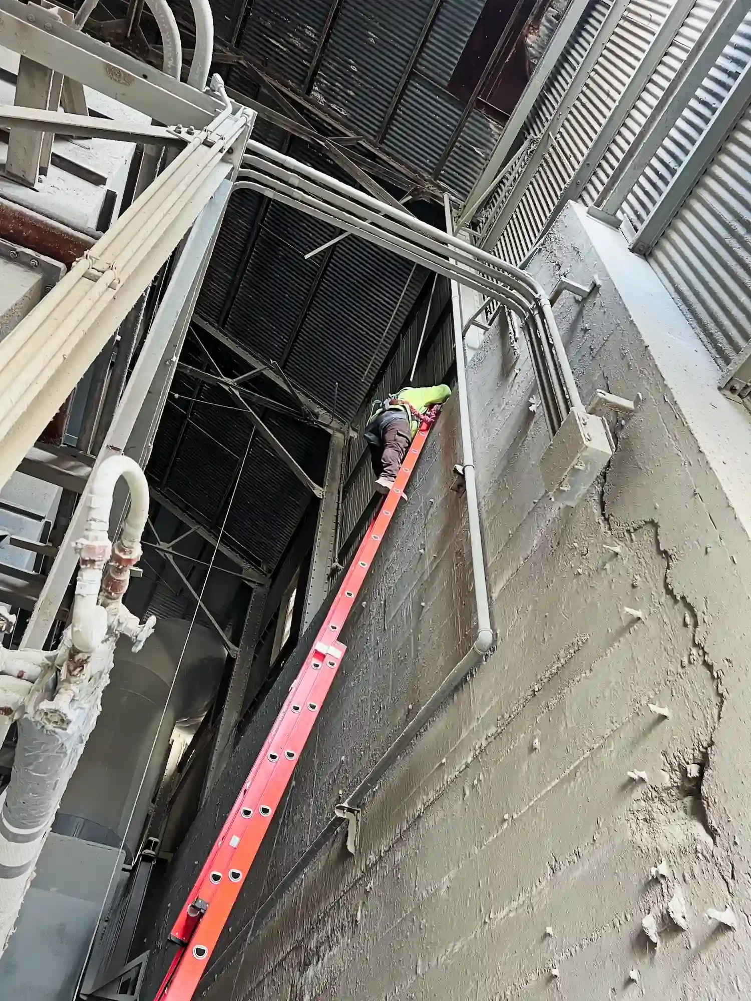 A worker is on a ladder inspecting or working on industrial equipment inside a building with exposed pipes and metal structures.