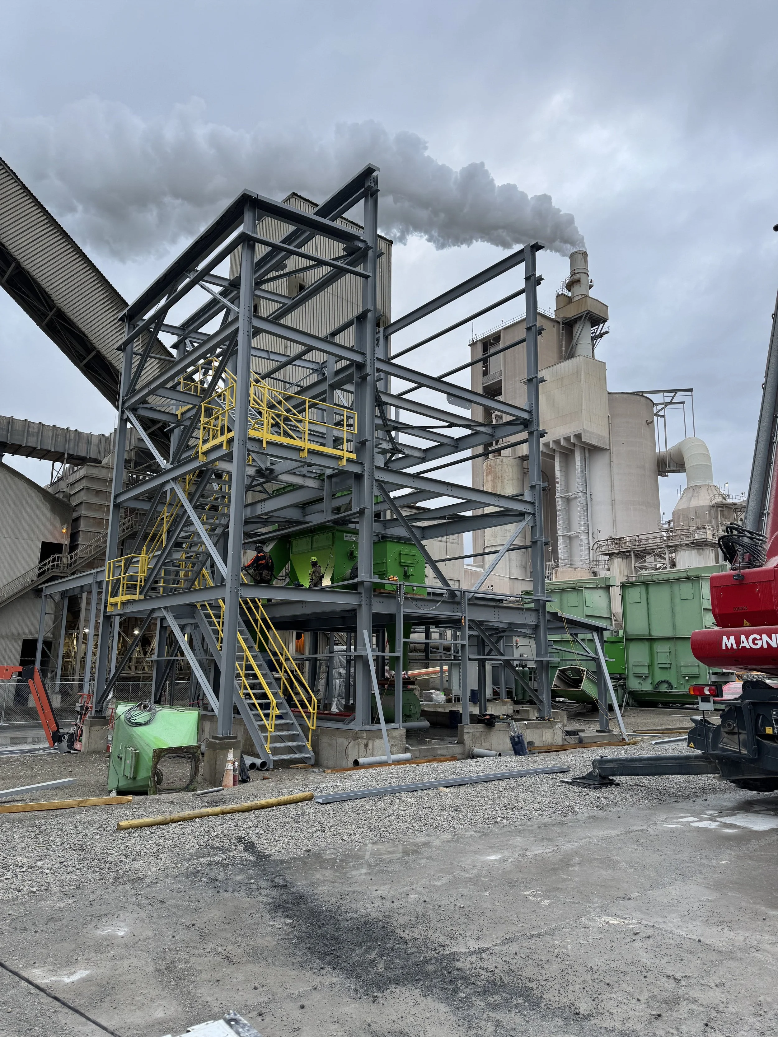 Industrial construction site with metal framework, workers, green equipment, and a factory emitting smoke in the background.