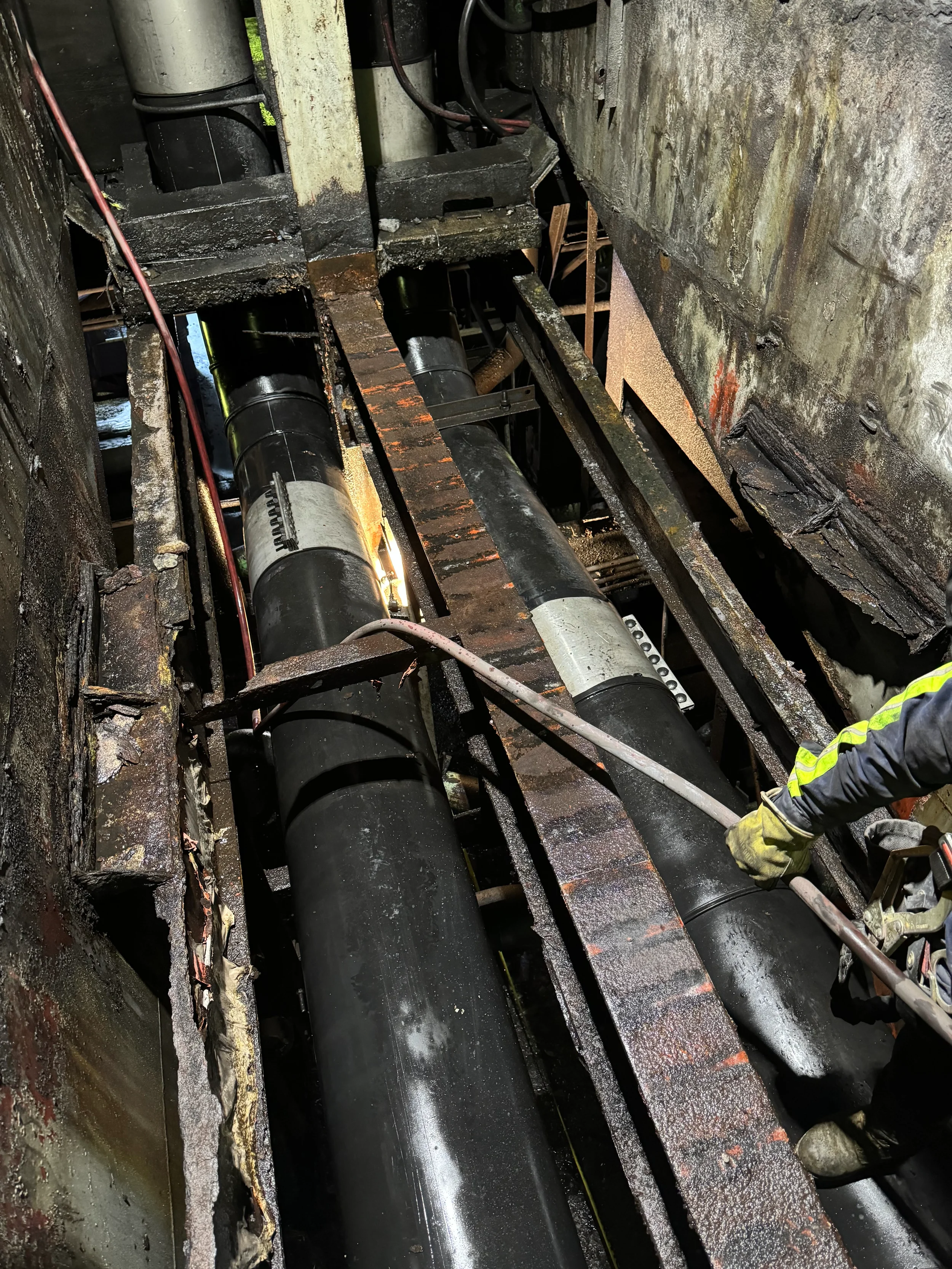 A worker inspecting large black pipes in a confined, corroded industrial space, with rust and grime on the surroundings.
