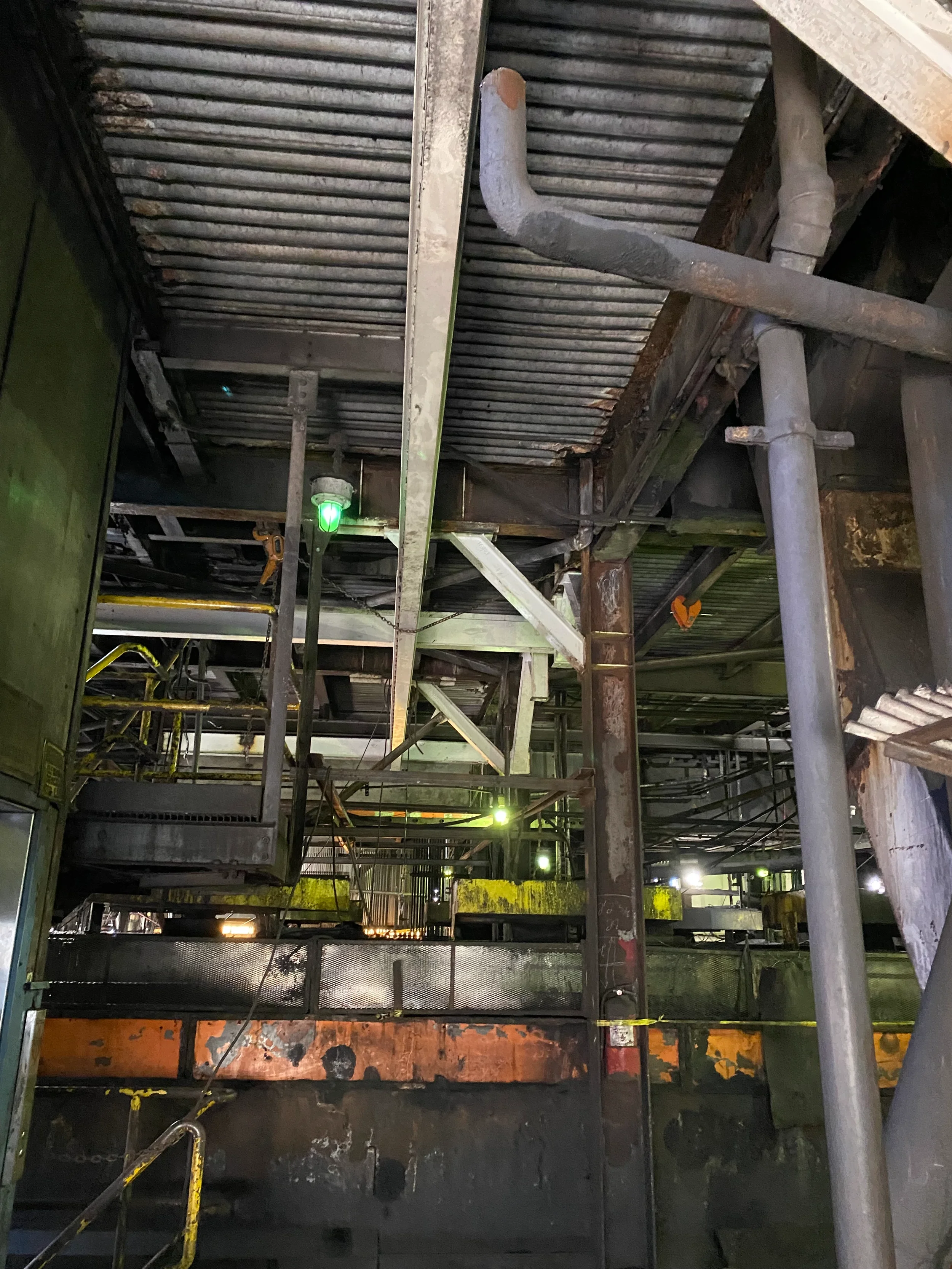 Industrial construction site with metal scaffolding, pipes, and temporary barriers, illuminated by green and yellow lights.