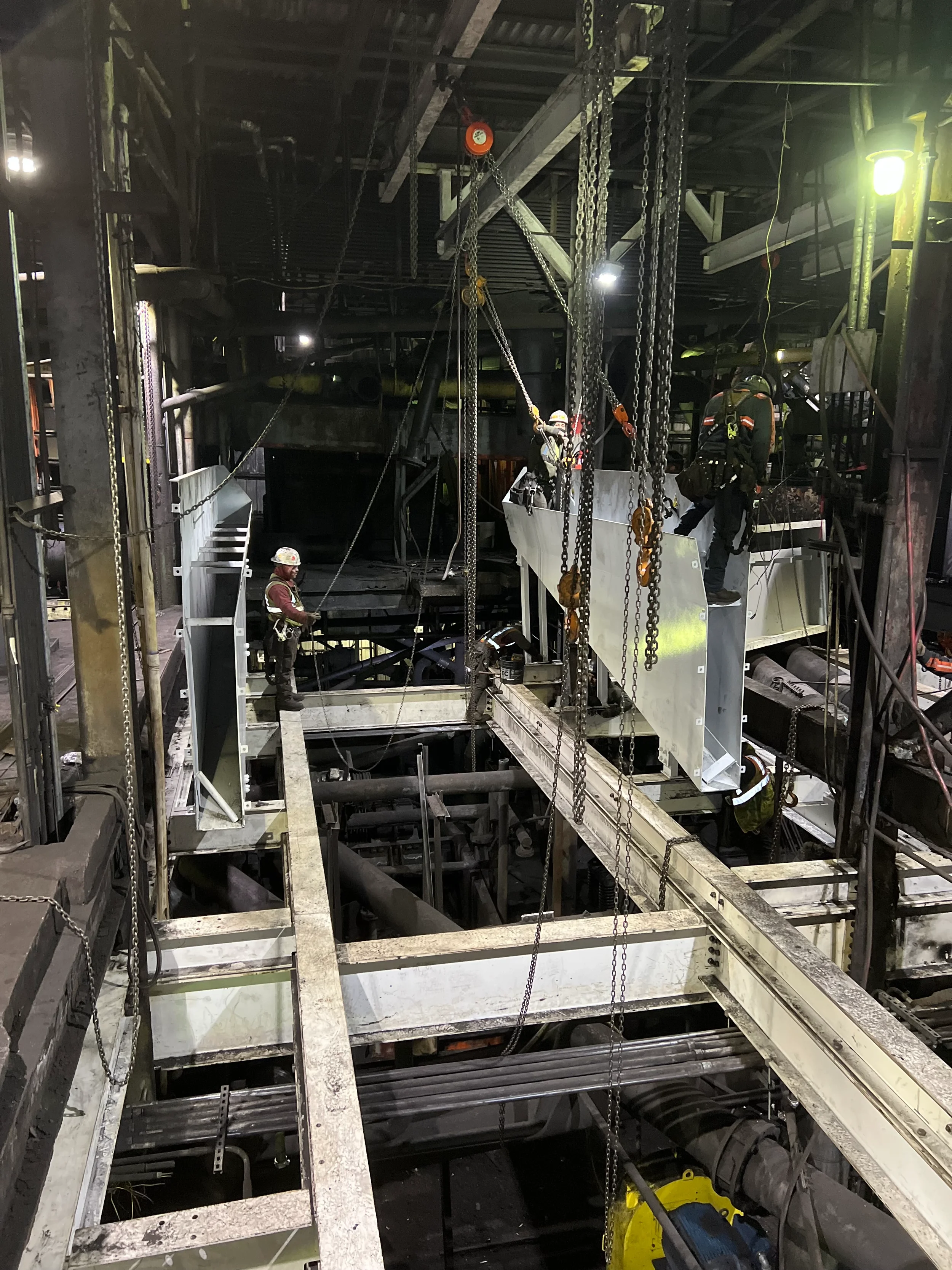 Workers in safety gear working on construction scaffolding inside a building, with pipes, chains, and structural metal framework visible.