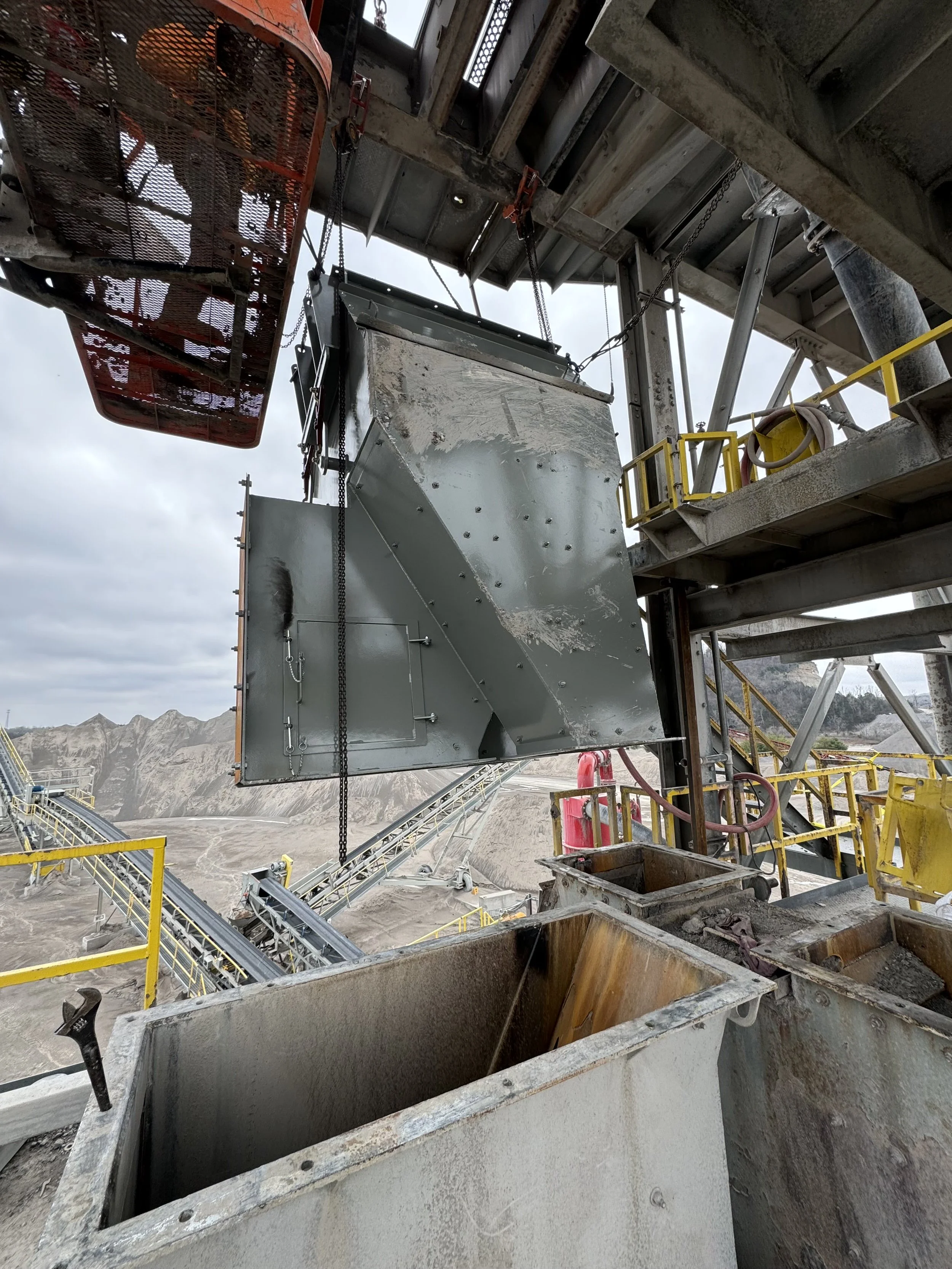 Construction site with metal structures, conveyor belts, and large industrial tags, with a mountainous background and cloudy sky.