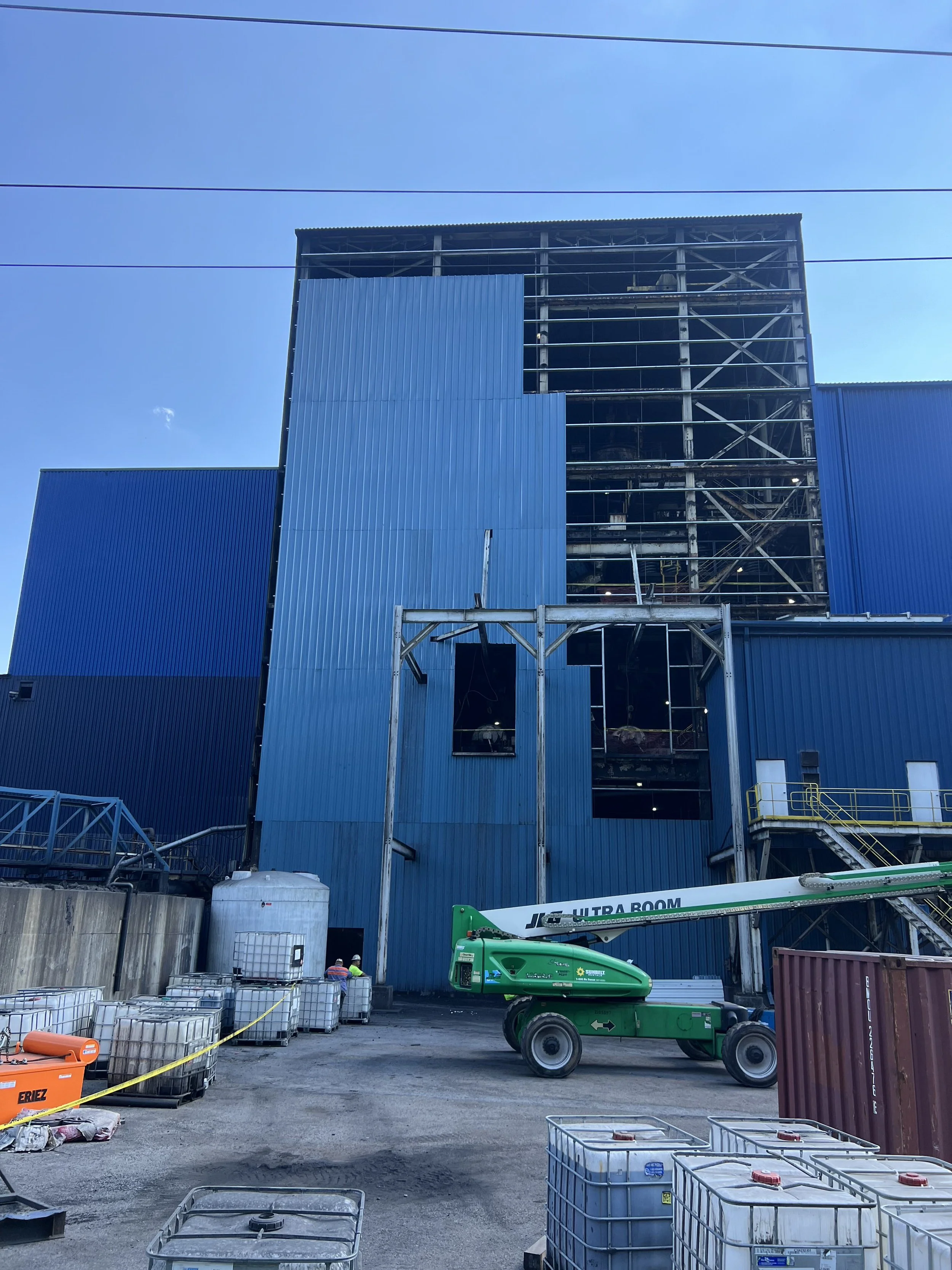 Construction site with a blue building under construction, a green lift, and several containers and construction materials in the foreground.