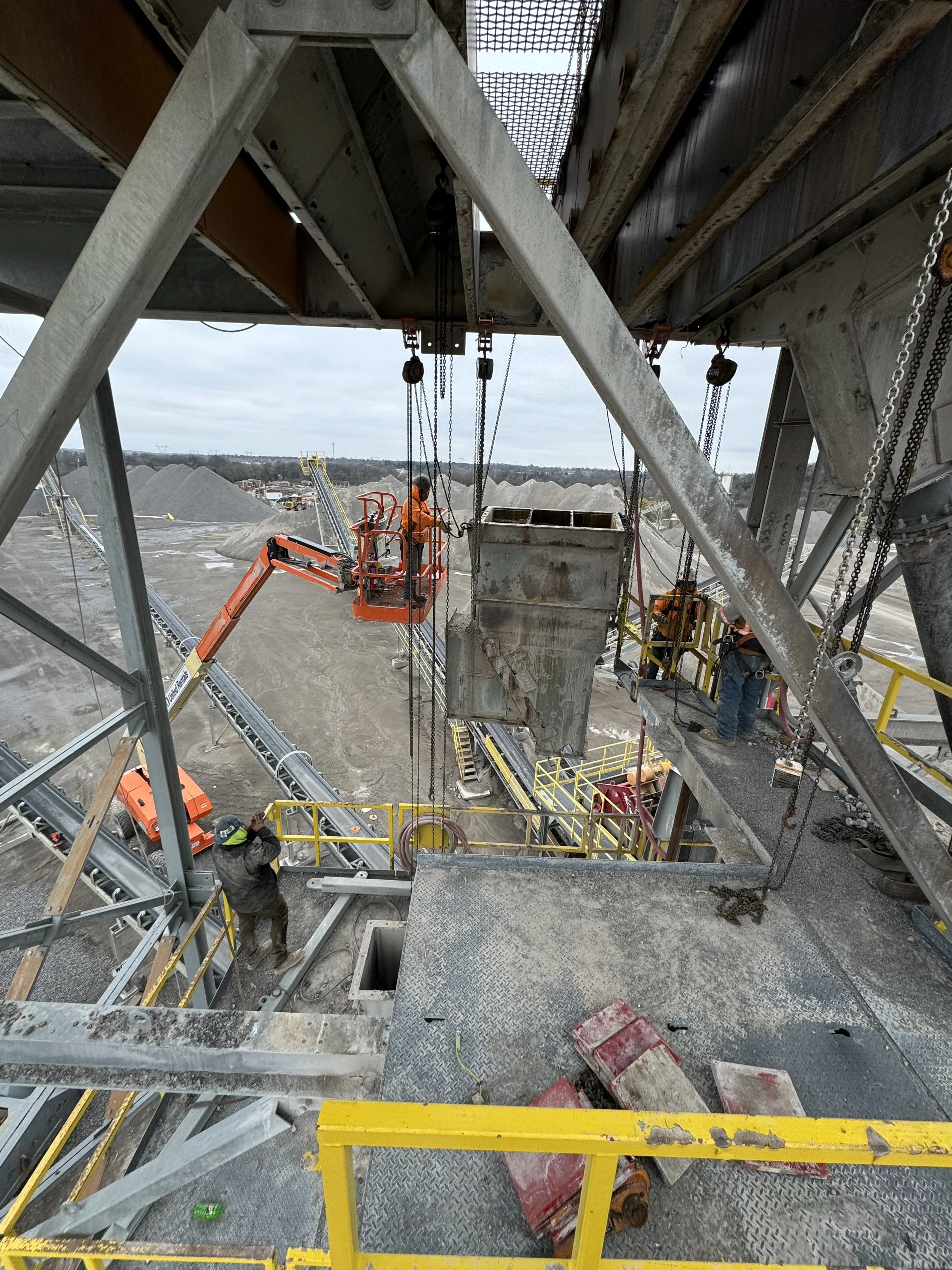 Workers operating on a construction site with large metal structures, chains, and equipment, with an overcast sky and mounds of gravel in the background.