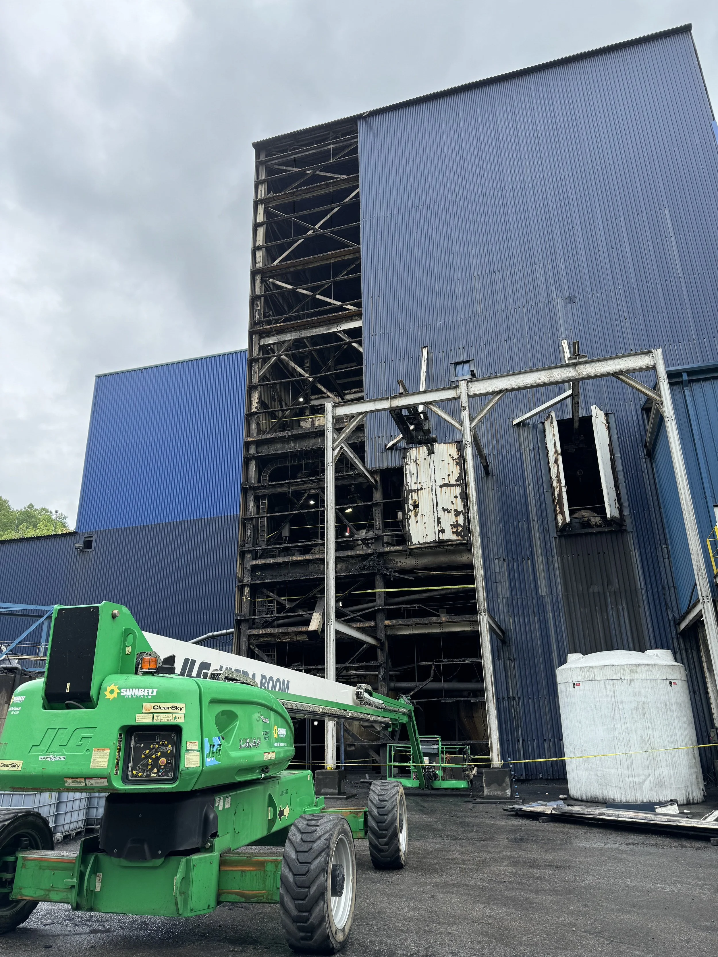 Construction site with a partially demolished blue building, metal framework, and a green lift in the foreground, under cloudy sky.