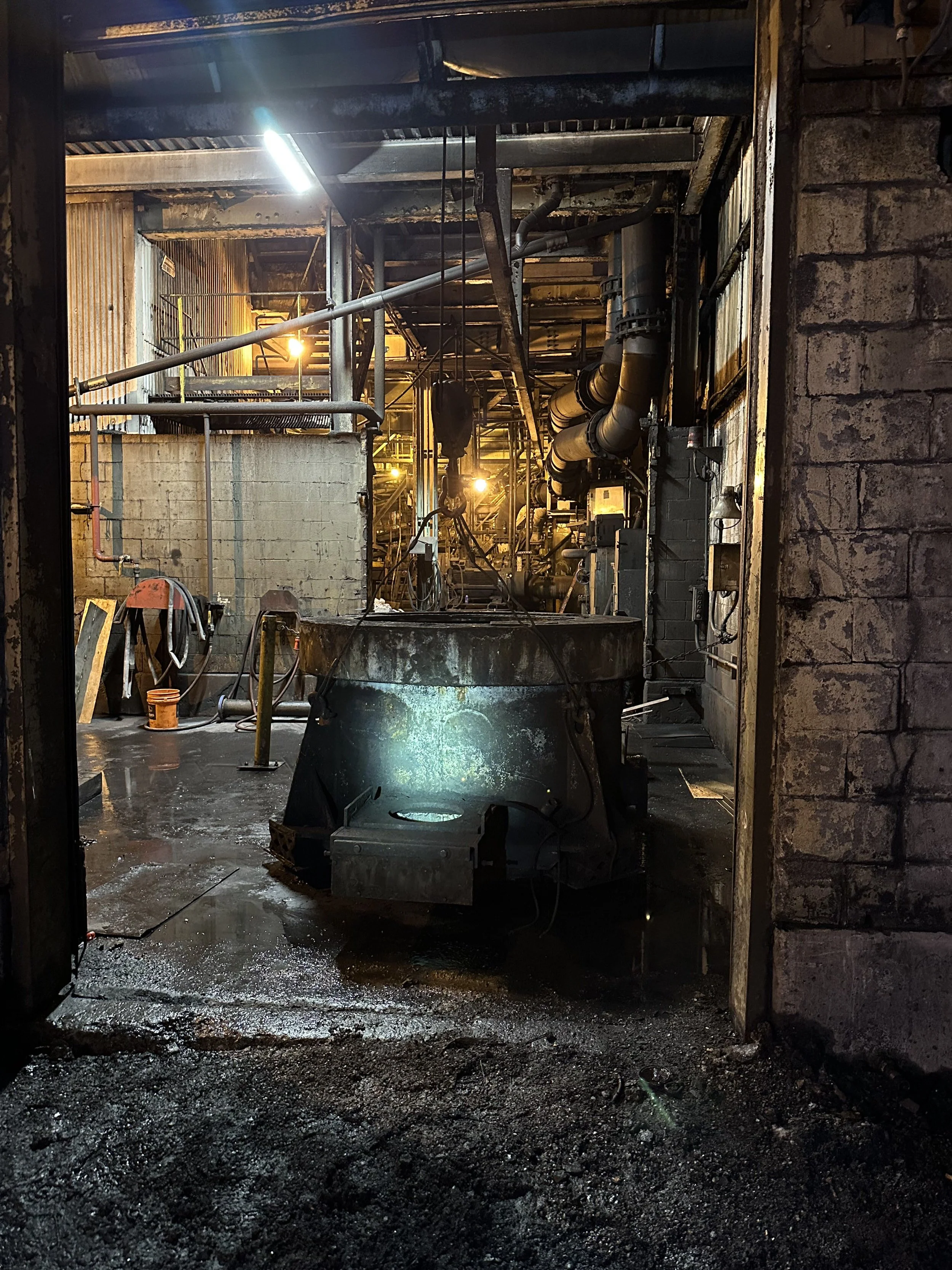 Industrial scene with pipes, scaffolding, and machinery, viewed through a doorway with brick and metal walls, wet floor with debris.