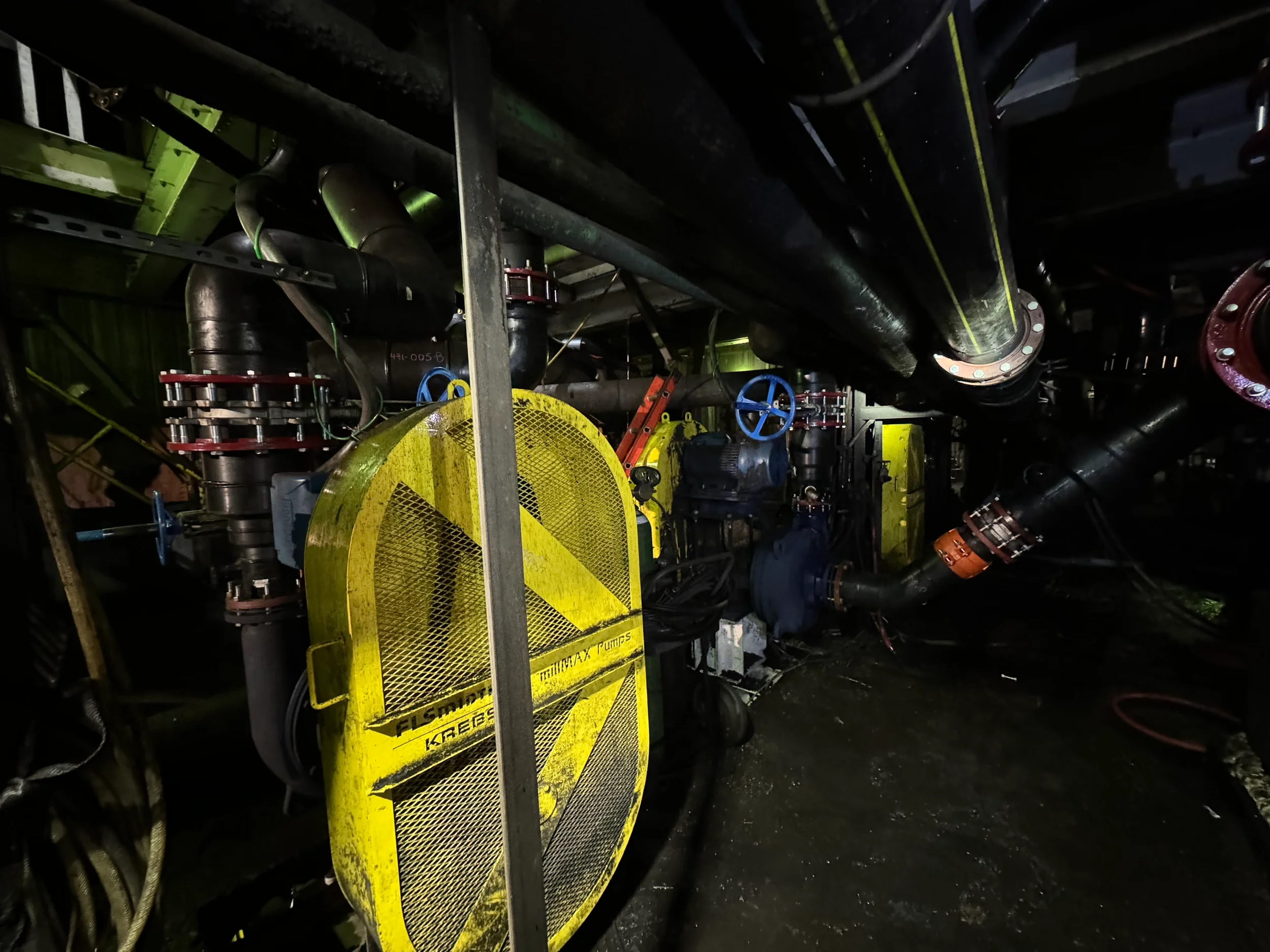An underground industrial setting with large black pipes, yellow safety equipment, and red and blue valves. The environment appears dark with minimal lighting.