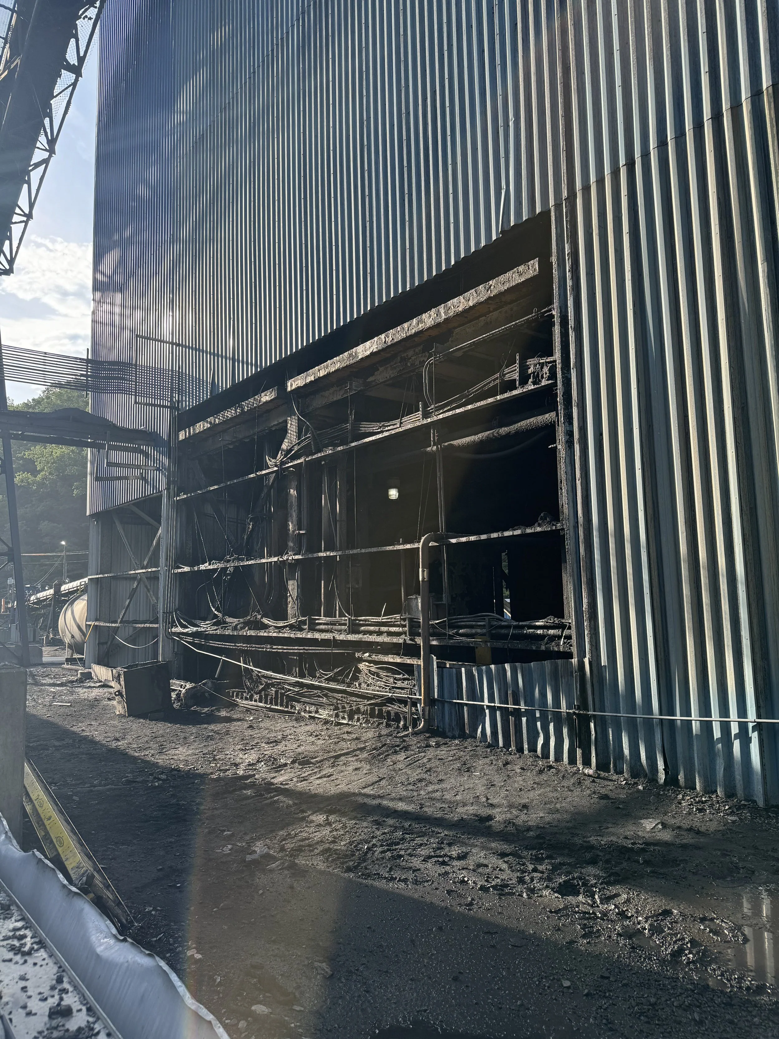 Building with grated metal siding and damaged exterior wall exposing interior wiring, with muddy ground and a barrier in the foreground.
