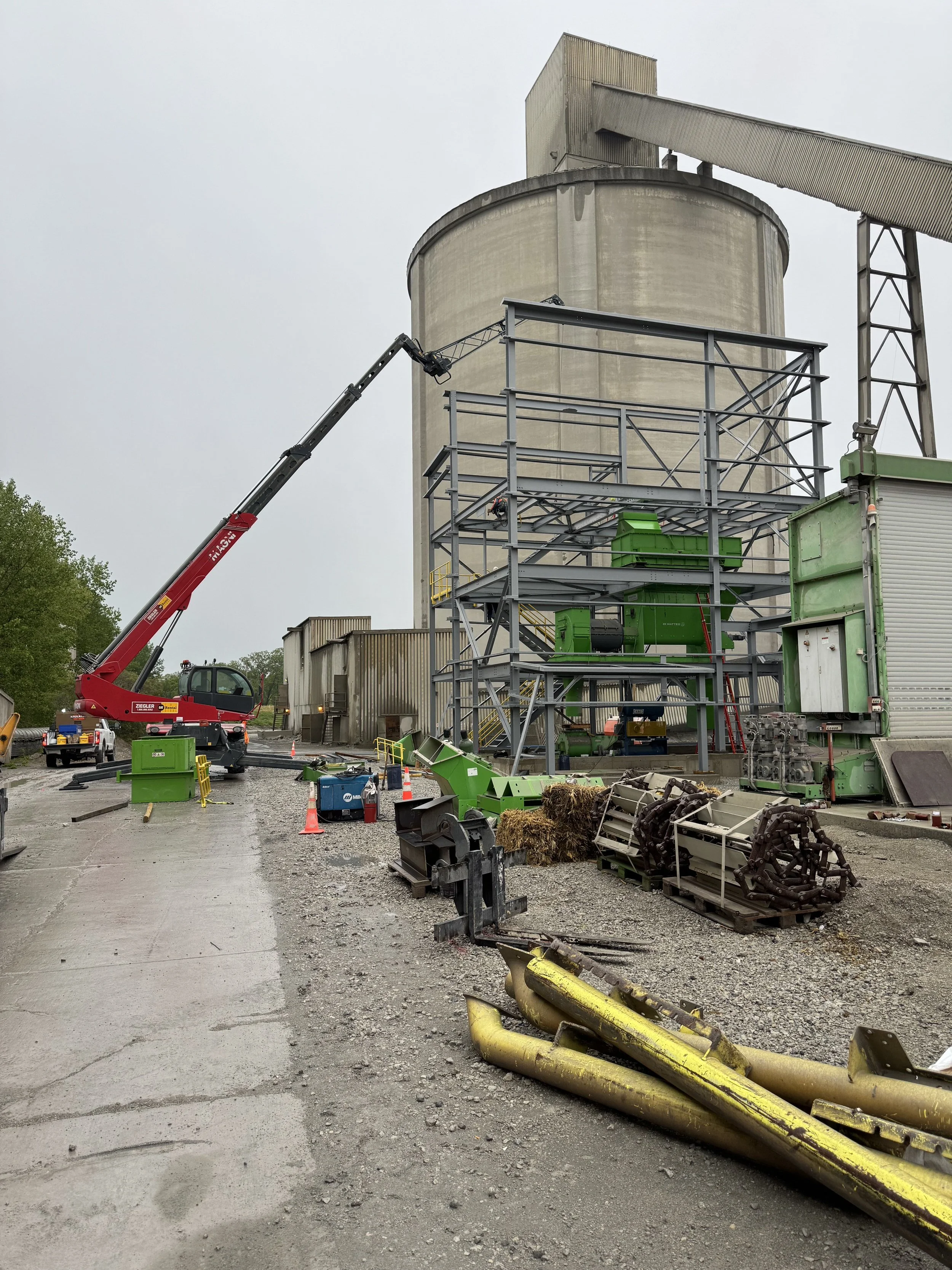 Construction site with scaffolding, a crane, and construction materials and debris on the ground, with a tall concrete silo in the background and a cloudy sky.