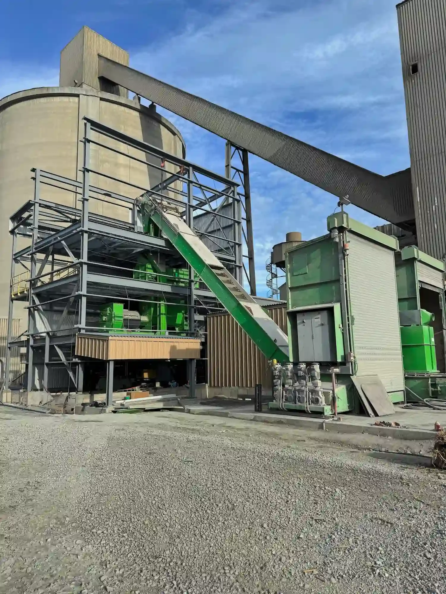 Industrial facility with large beige silo, green machinery, and metal structures, under a blue sky with some clouds.
