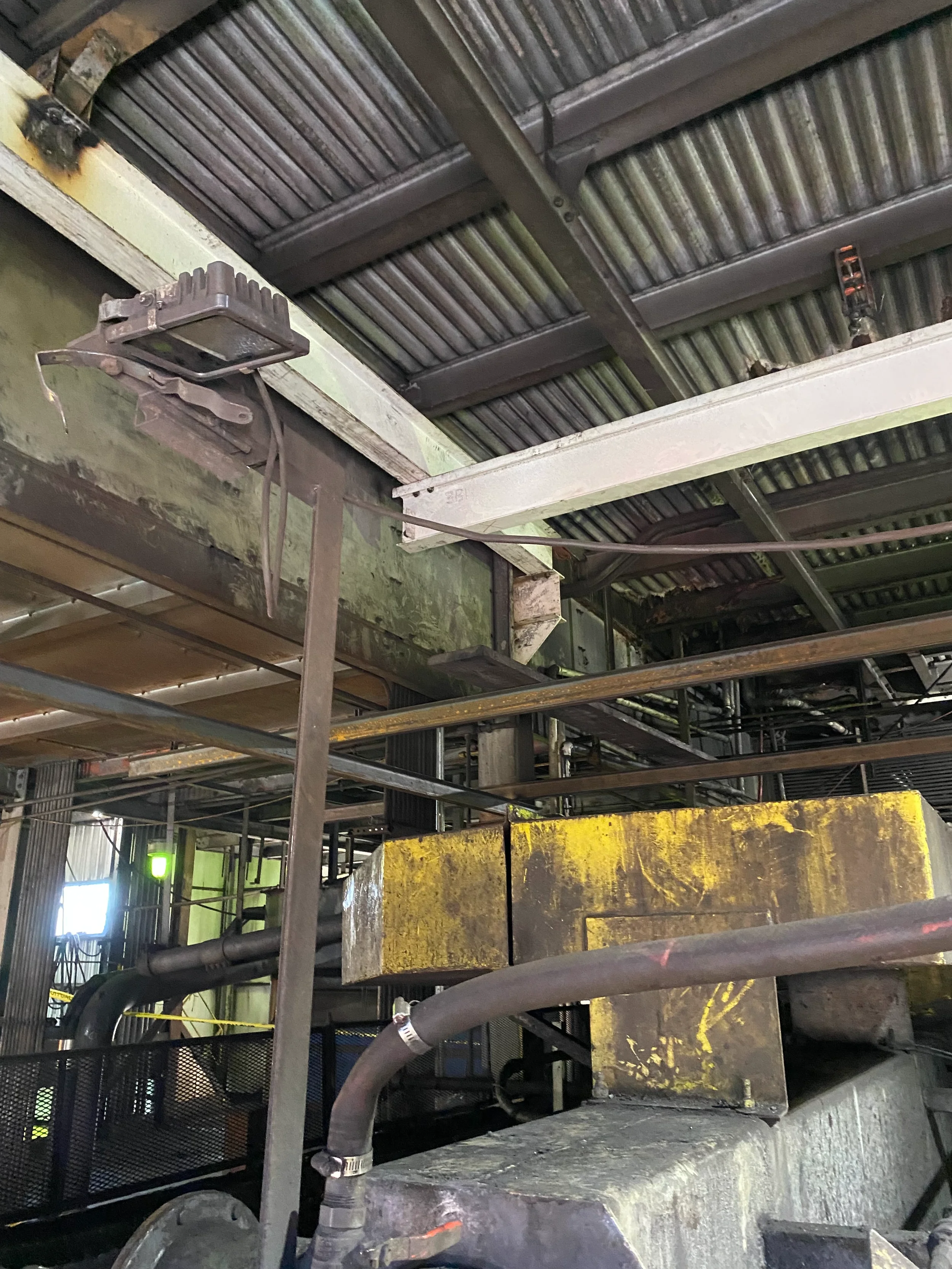 Industrial interior with metal pipes, a large yellow machine part, and a metal framework overhead.