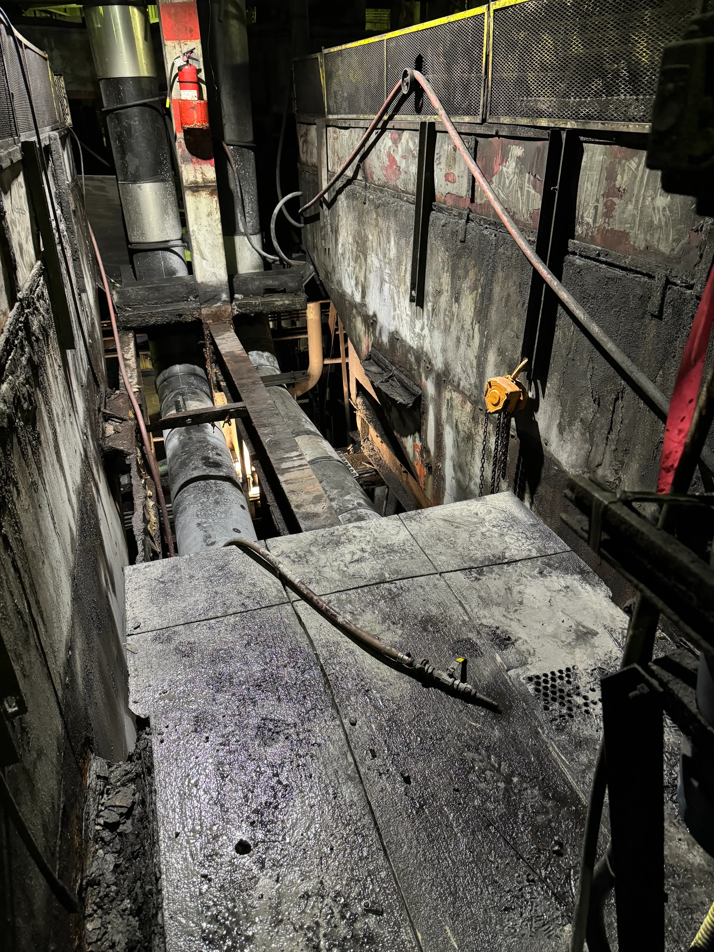 Damaged industrial setting with charred walls, wet and burnt floor, pipes, and fire extinguisher, indicating fire damage.