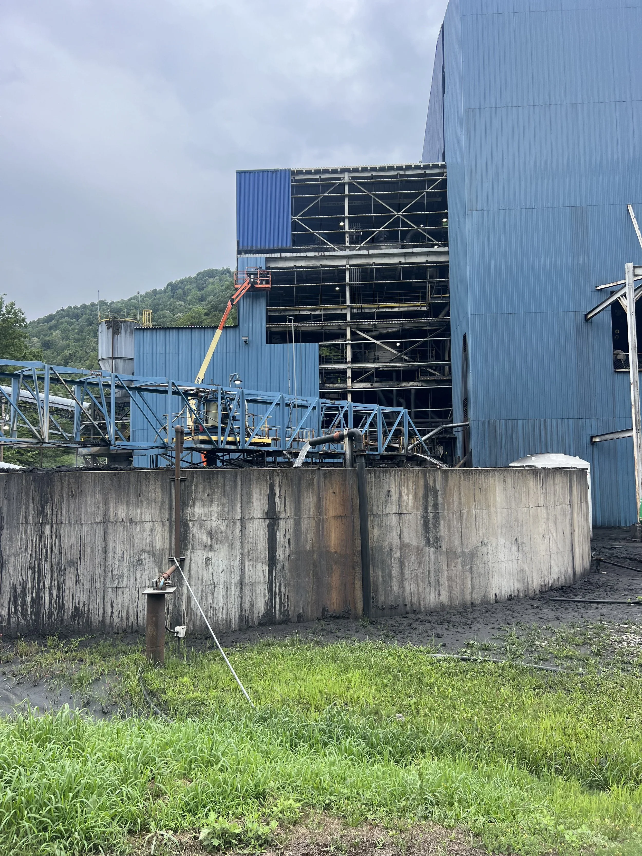 Exterior of an industrial building with blue corrugated metal walls and scaffolding, overgrown grass in the foreground, and hills in the background under a cloudy sky.