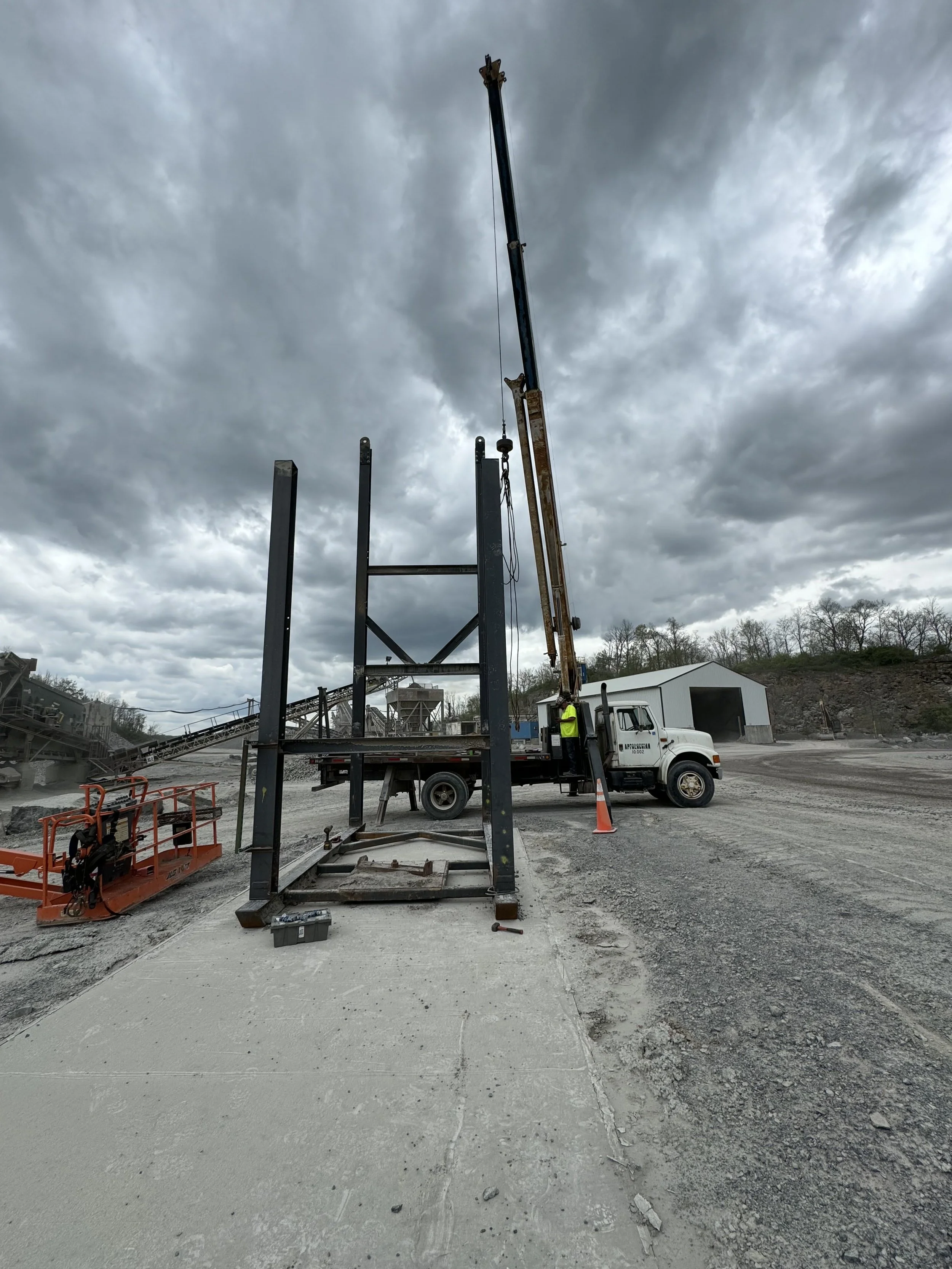 Construction site with a crane lifting through a steel frame structure and dark cloudy sky overhead.
