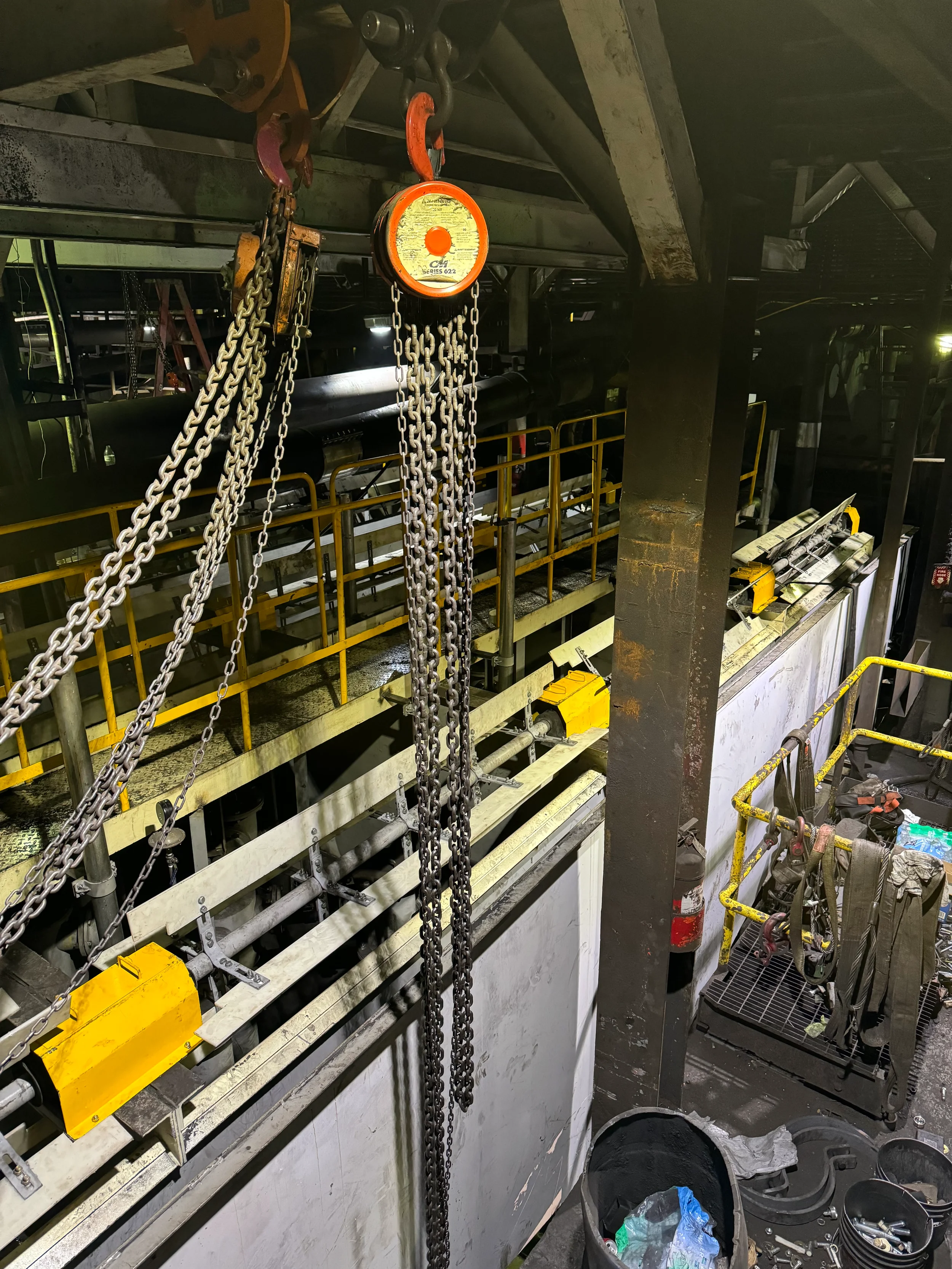 Industrial workspace with chains hanging from a hoist, yellow safety barriers, tools and equipment scattered around, and a dark environment.